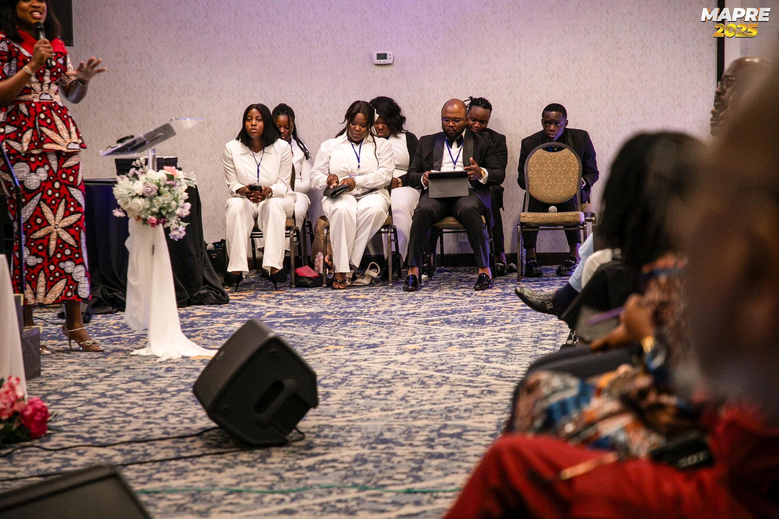 A woman in a colorful patterned dress speaks into a microphone while gesturing with her hands, standing near a table with a floral arrangement. Behind her, a group of people, mostly dressed in white and dark suits, are seated on chairs in a conference or event room.
