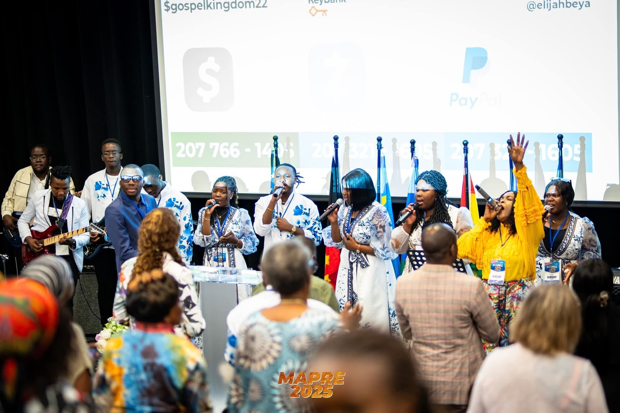 Group of people singing and playing guitar on stage at a conference or event, with an audience in the foreground. A large screen in the background displays text and social media handles.
