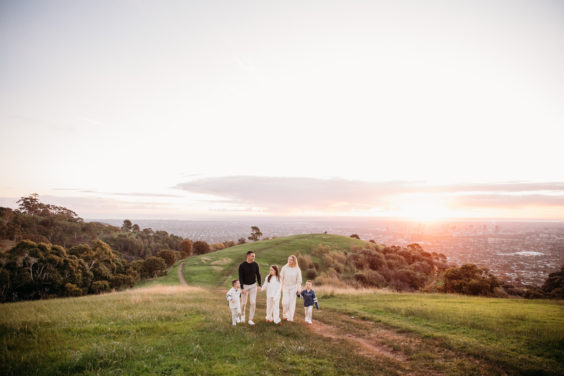 adelaide-fFamily of five walks hand in hand across rolling hills overlooking Adelaide during a sunset family photography session.amily-photographer-family-walking-hills