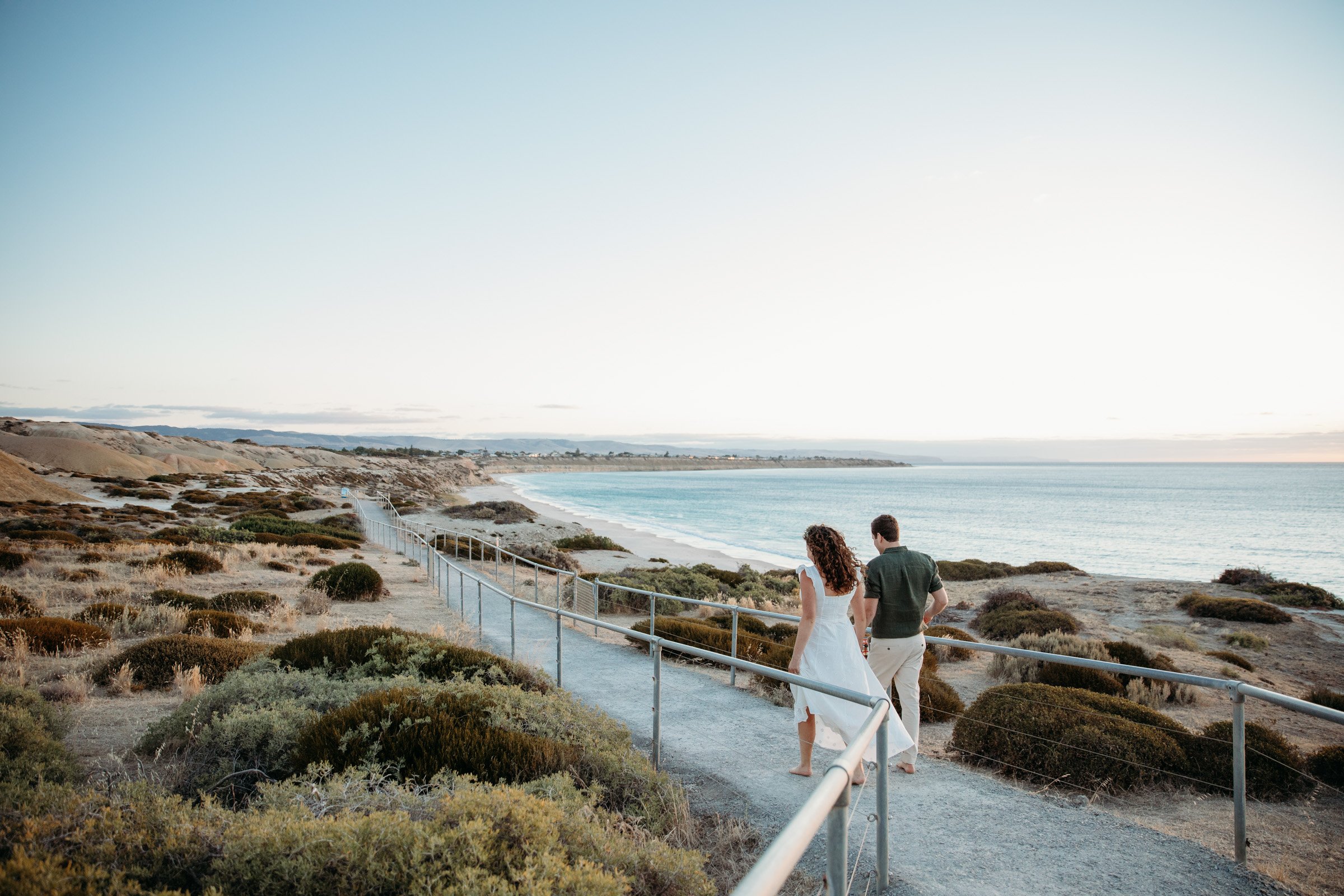 A couple walking along a seaside boardwalk with coastal vegetation overlooking the ocean under a clear sky.