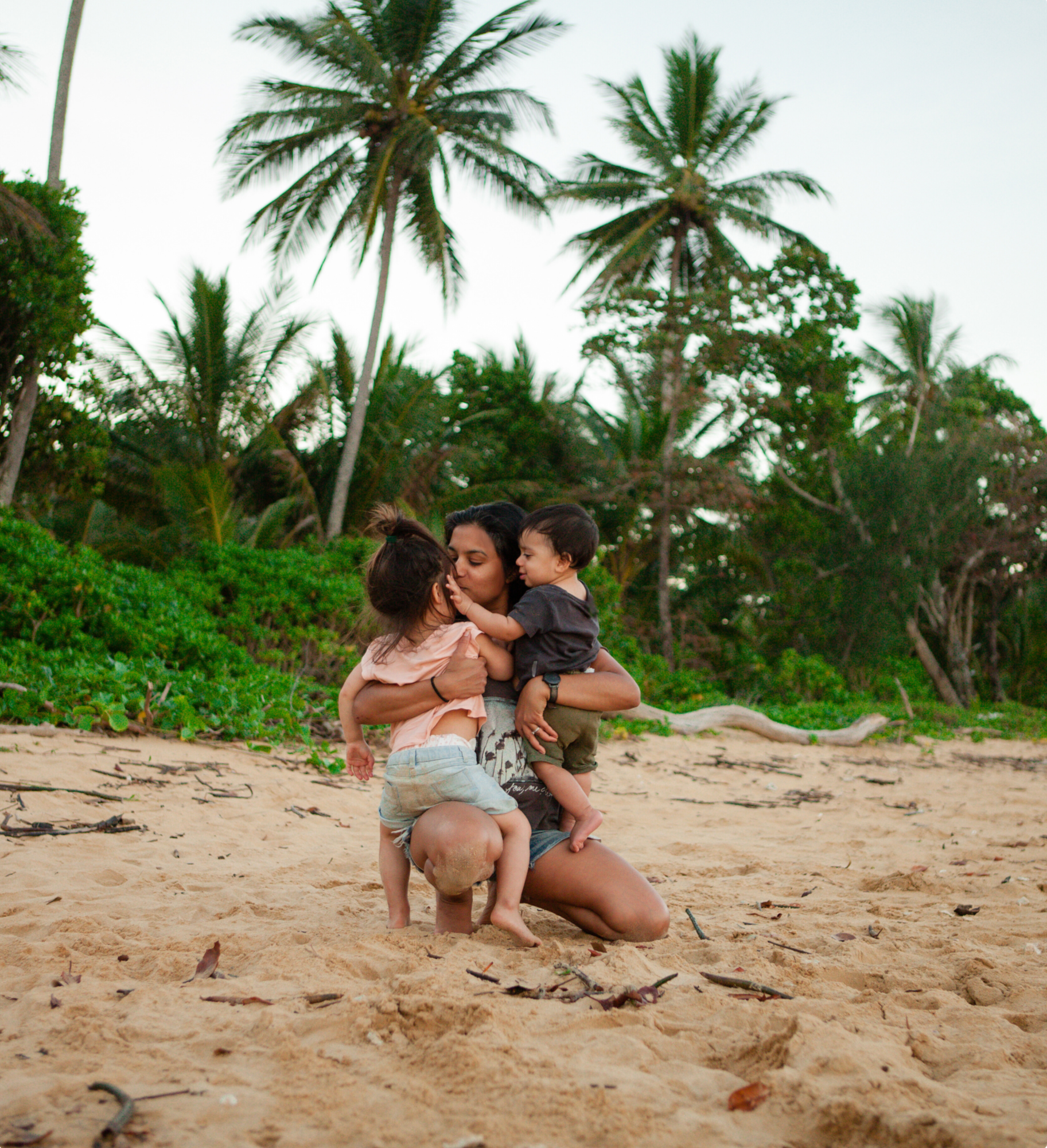 Photographer cuddles two young children on a sandy beach surrounded by tropical palm trees, reflecting a warm, natural and adventure-filled approach to family photography.