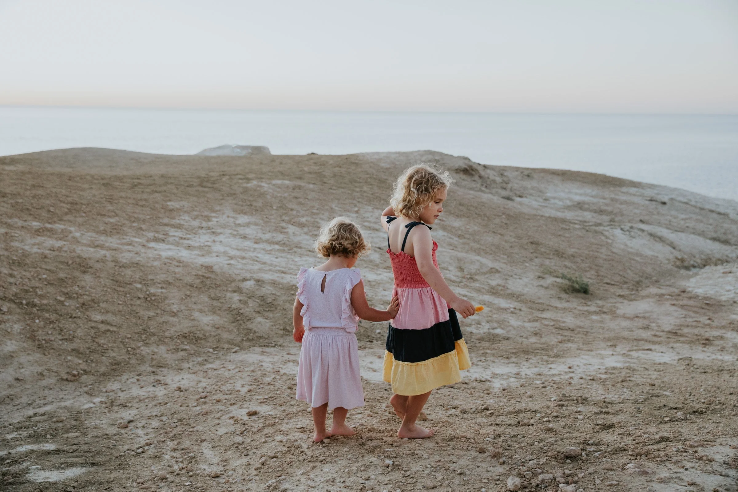 Two young sisters holding hands as they walk along a coastal path overlooking the ocean.