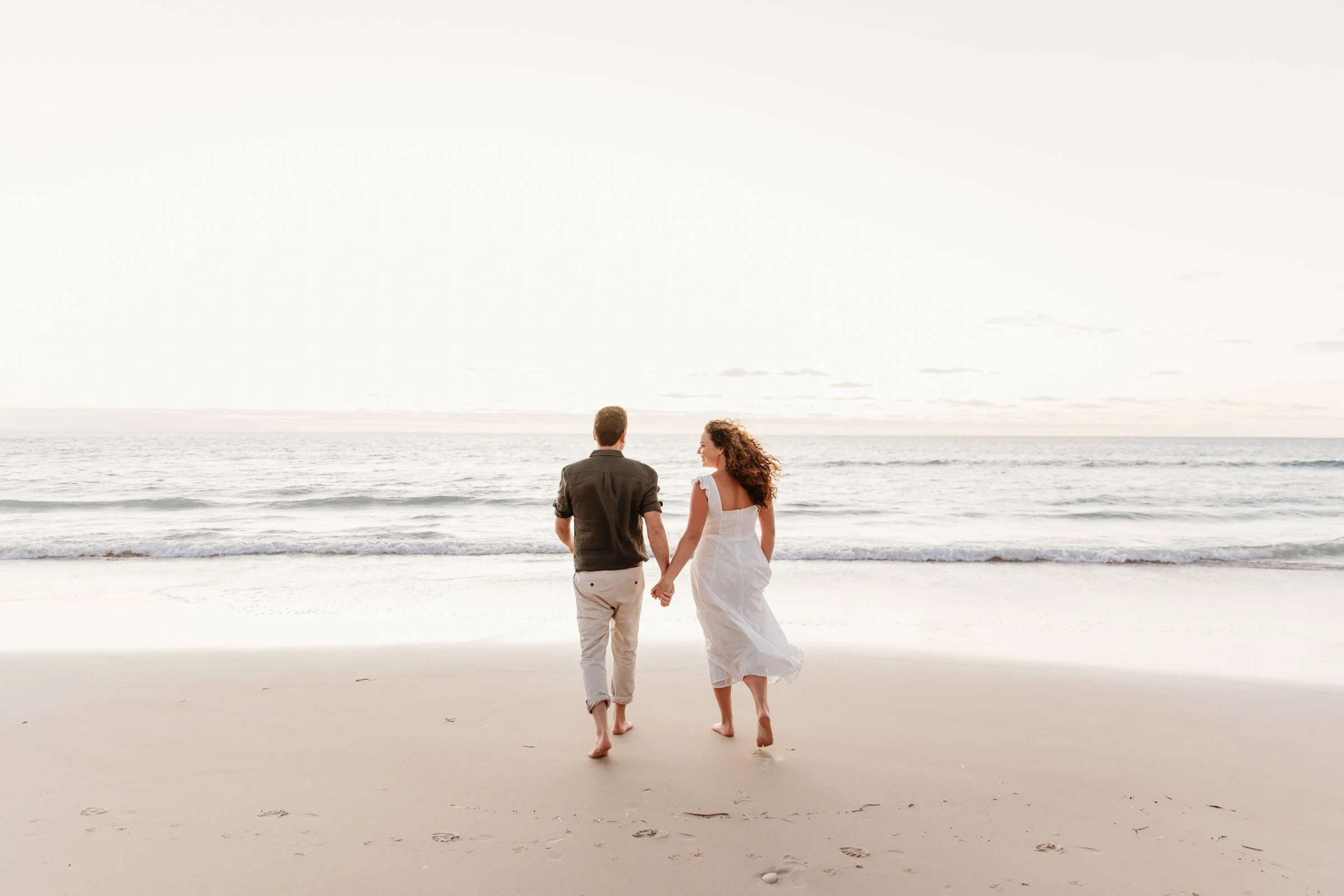 A couple walking hand in hand along the beach at sunset, with the ocean and sky in the background.