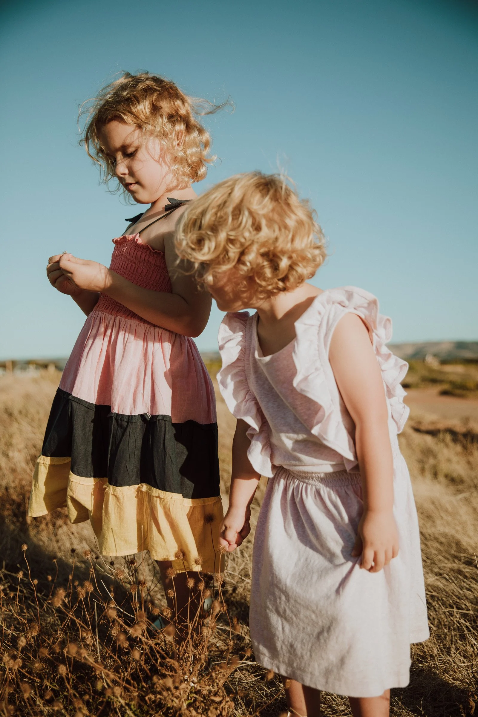 Two young girls with curly blond hair walking hand in hand outdoors in a field with dry grass, under a clear blue sky.