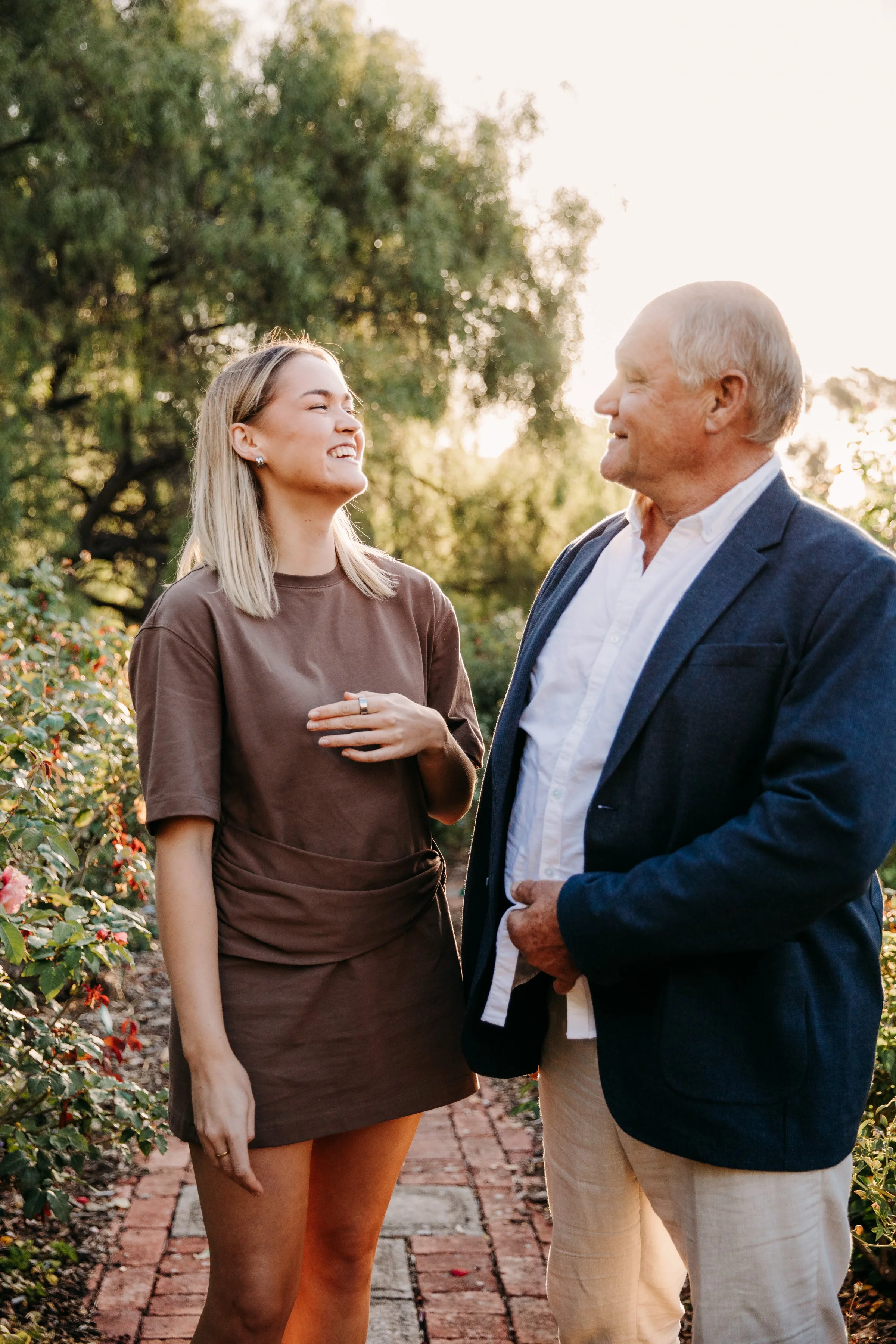 A young woman and an older man are standing outdoors on a brick pathway, smiling at each other. The woman is wearing a brown dress and the man is dressed in a dark blazer, white shirt, and beige pants. There are green trees and bushes in the background, and sunlight is shining through the trees.