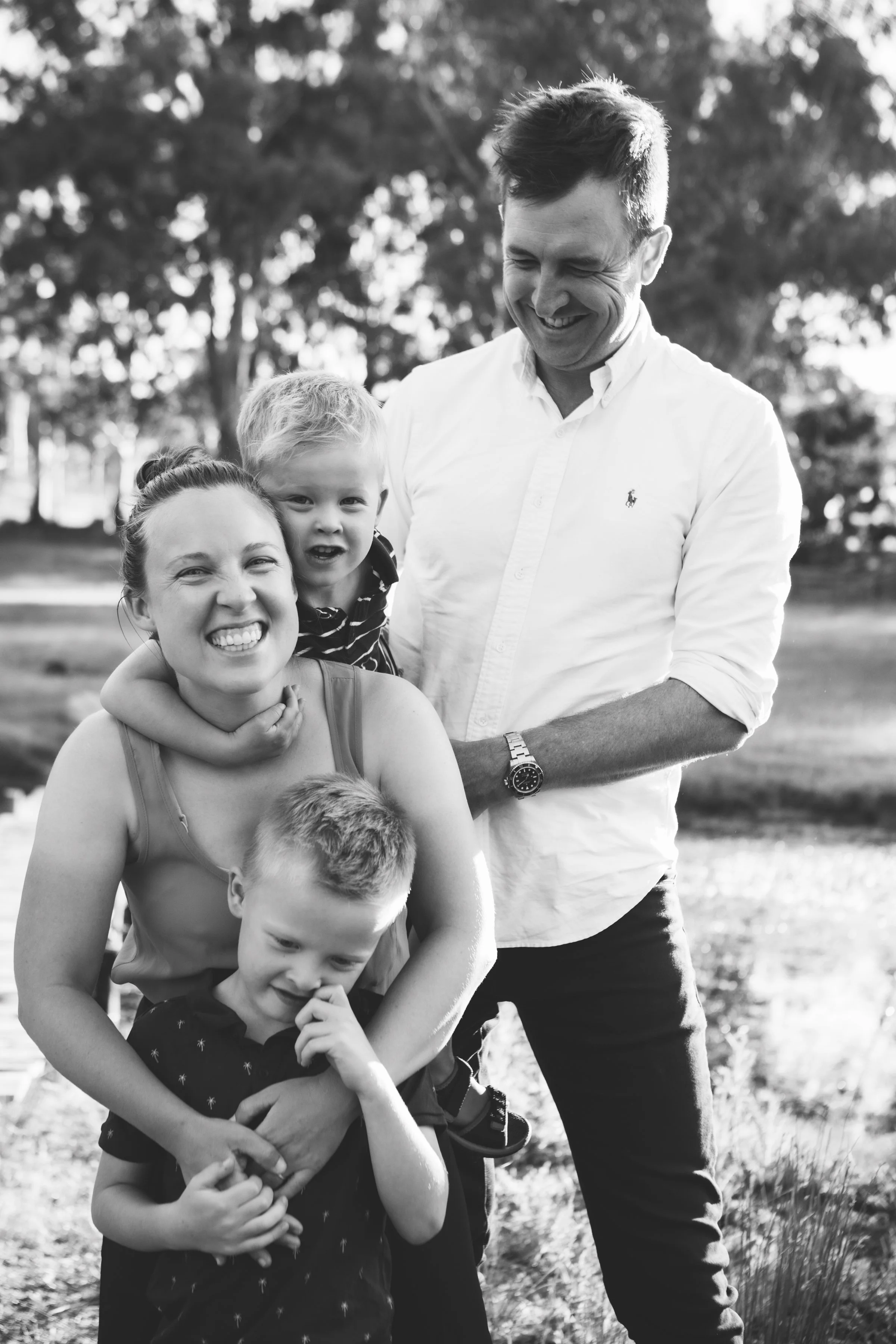 A smiling woman with her eyes closed, two young boys, and a man outdoors in a park with trees in the background.
