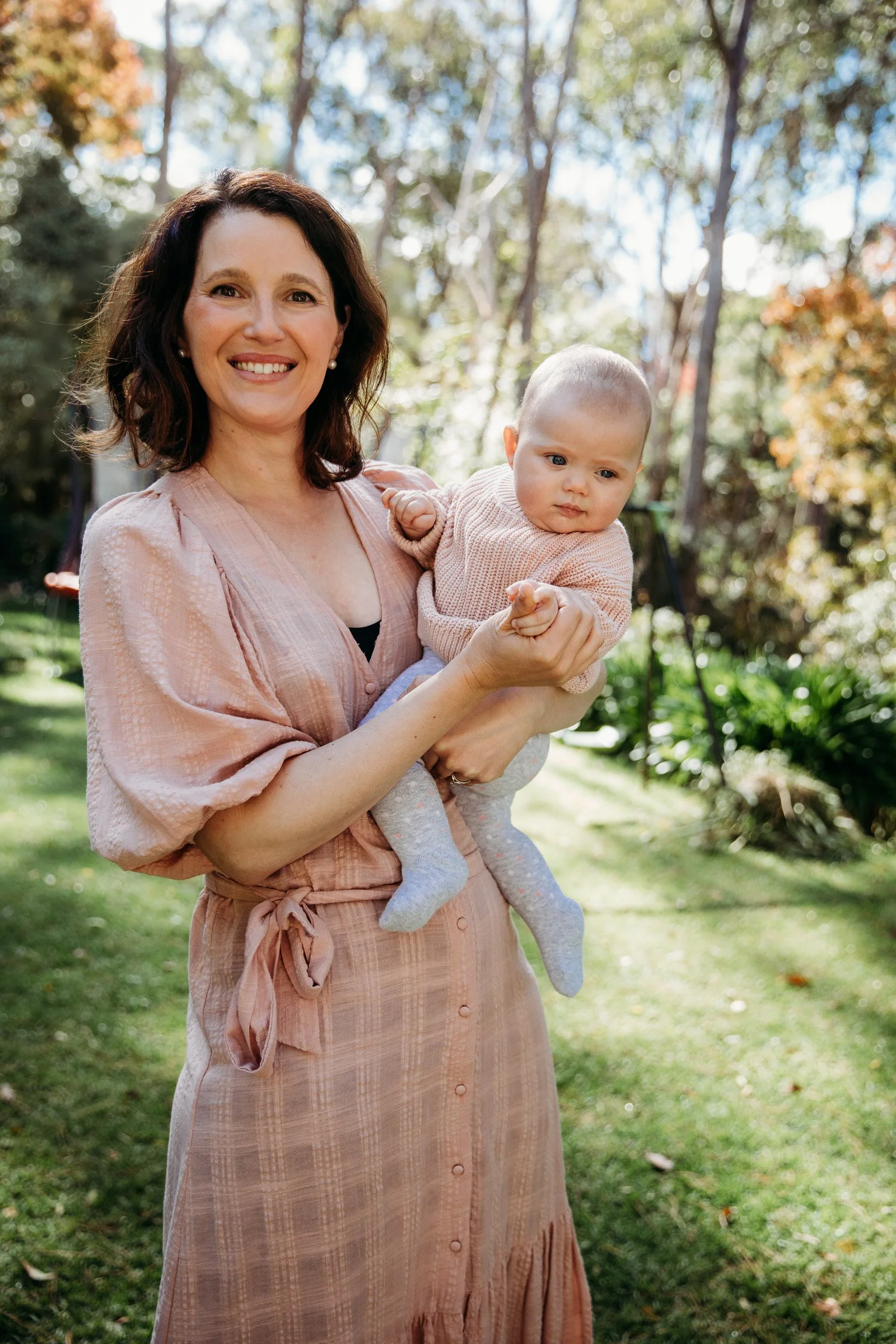 A woman with shoulder-length dark hair holding a baby in an outdoor park with trees and grass, during daytime.