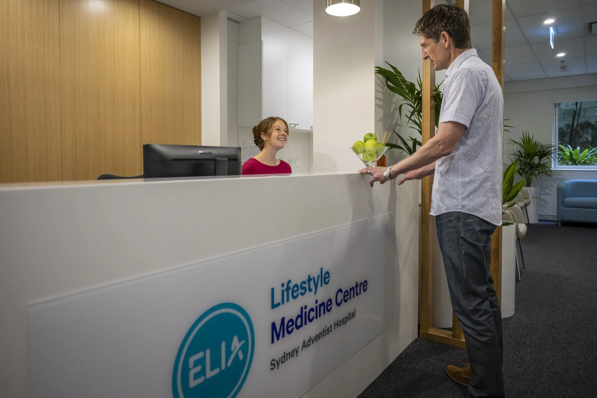 A man is handing a bowl of apples to a woman at a reception desk in a medical center, with plants and seating in the background.