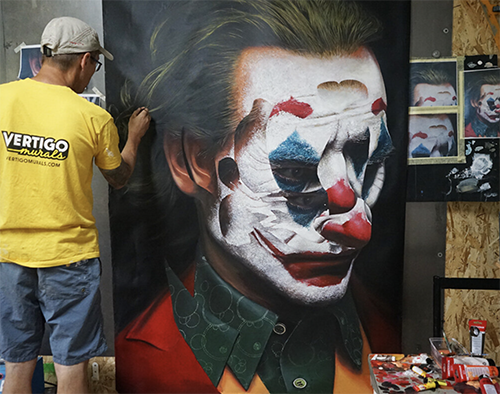 Artist in a yellow shirt working on a large portrait of a person with clown makeup, styled like the Joker, with bright face paint, green hair, and a red jacket.