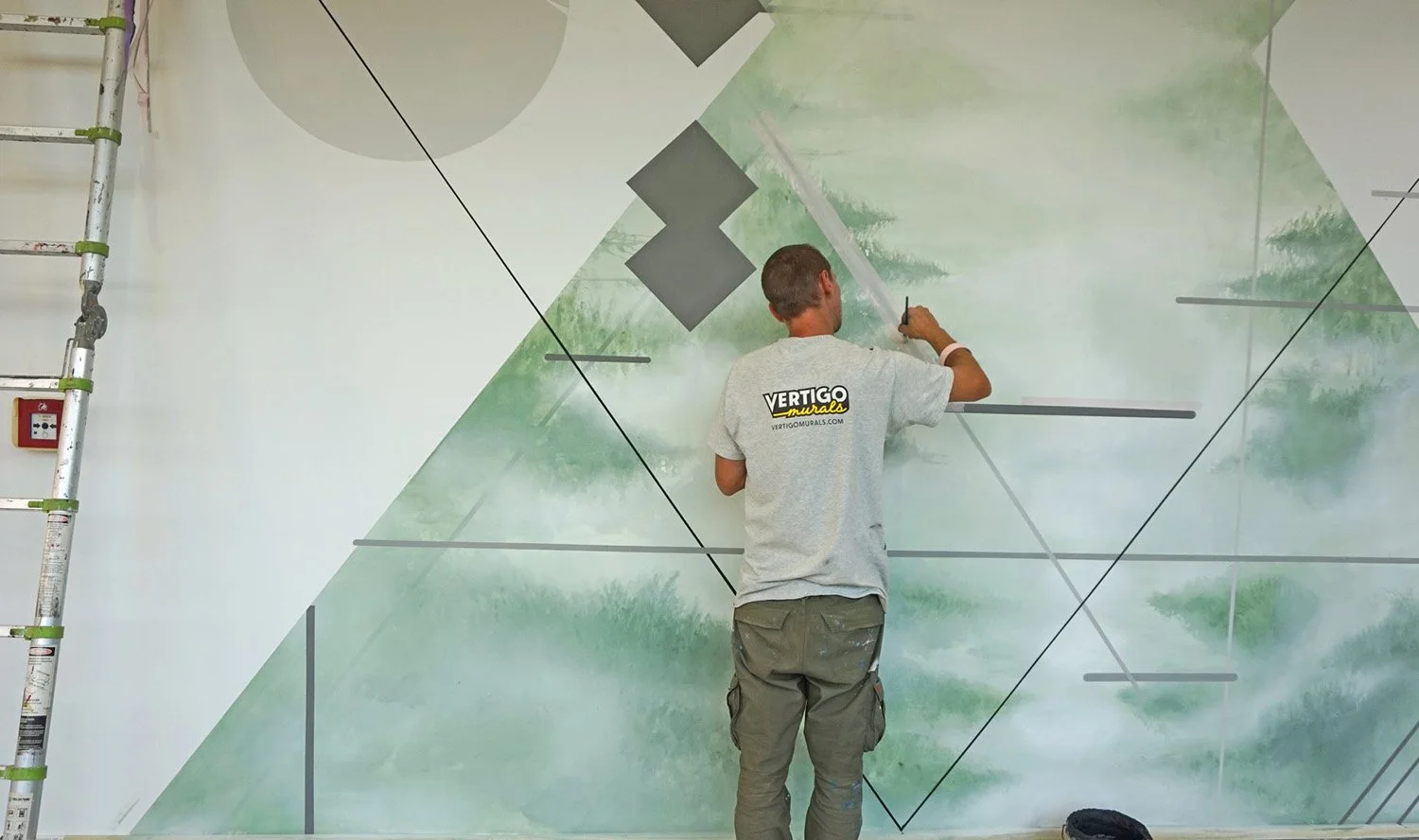 A man painting a mural on a wall with geometric shapes and a nature scene, wearing a gray T-shirt with company branding and khaki cargo shorts, standing on the floor near a step ladder.