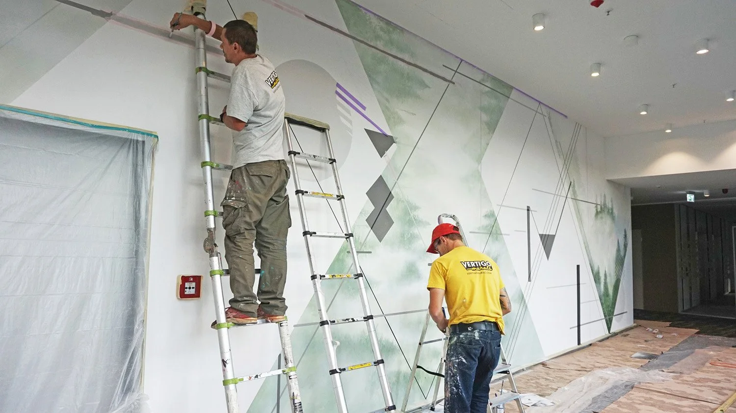 Two workers painting a mural on a white wall inside a building, using ladders for access.