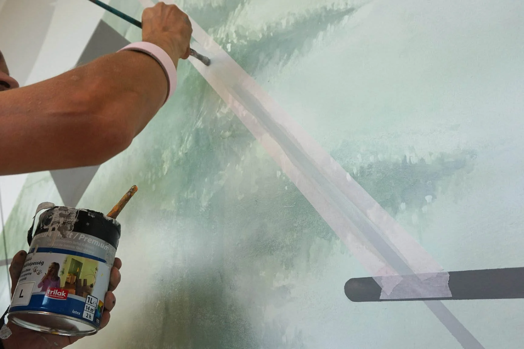 A person painting a wall with a brush, using a container of paint in their left hand, with masking tape covering part of the wall.