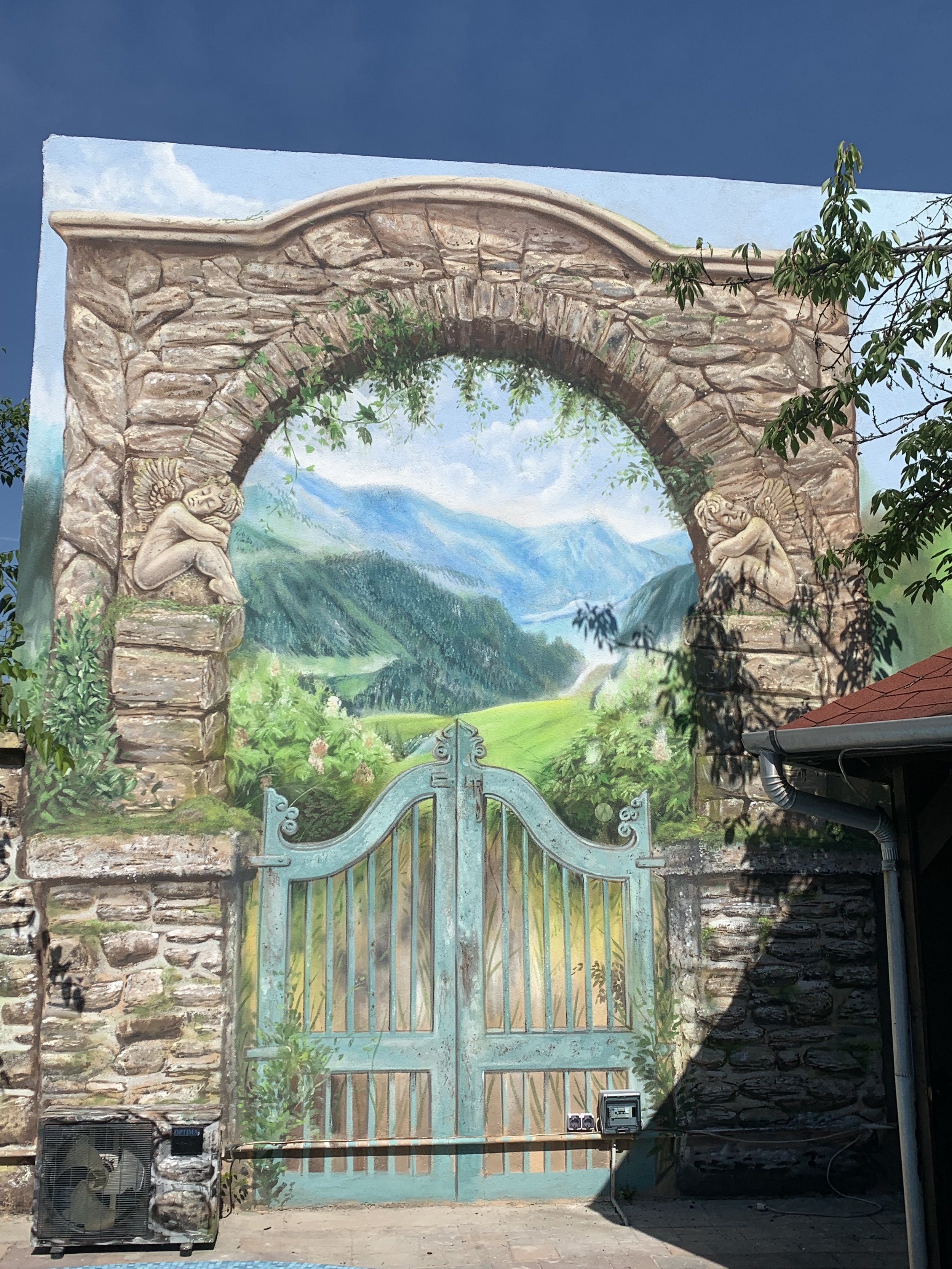 A mural depicting an arched stone gate with two sculptures of sleeping faeries on either side, a scenic landscape of mountains, forests, and a blue sky painted behind the gate, with some tree branches and shadows in the foreground.