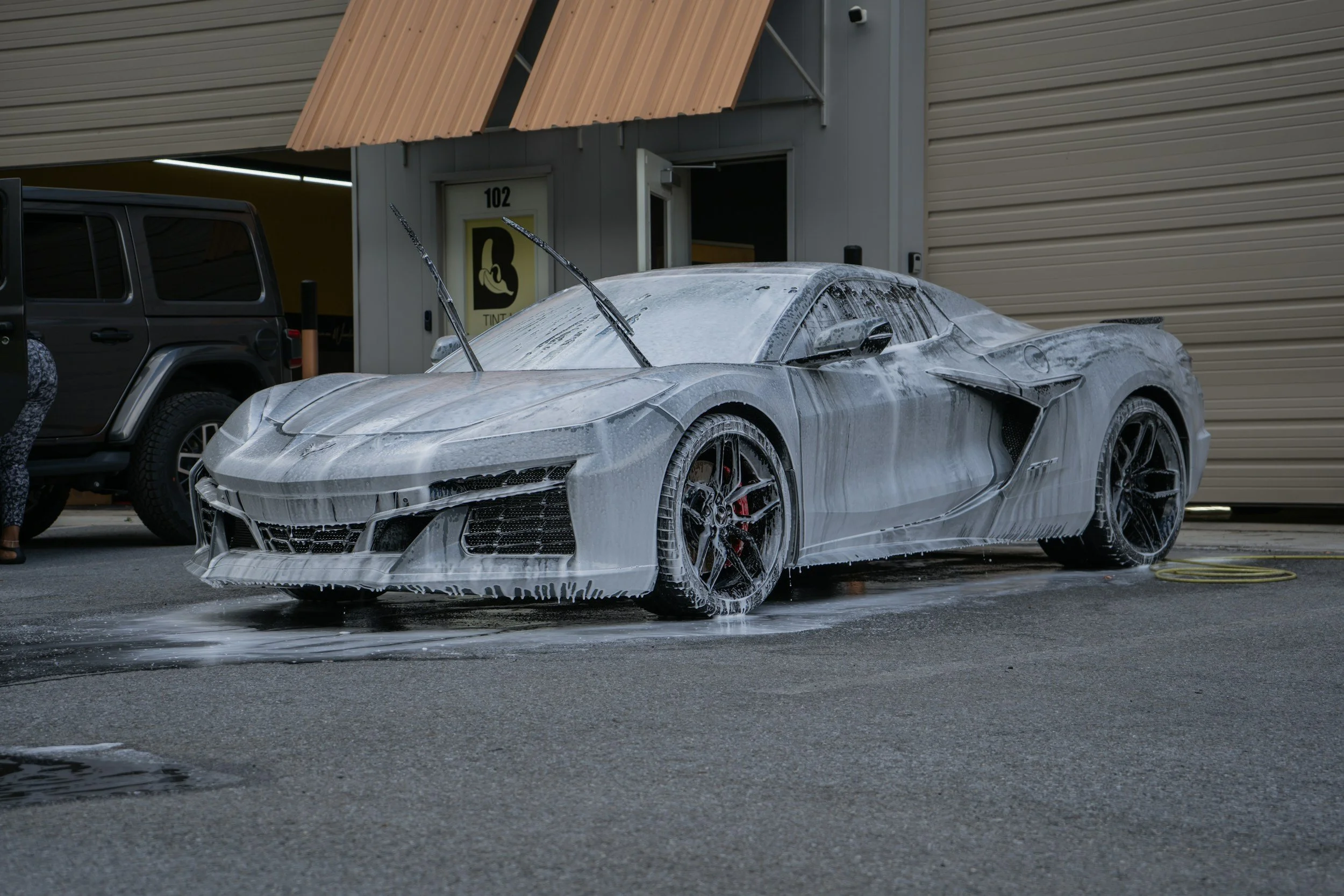 A silver sports car covered in soap foam during a car wash, with soap dripping down the sides, parked outside a building with a roller door and a yellow sign.
