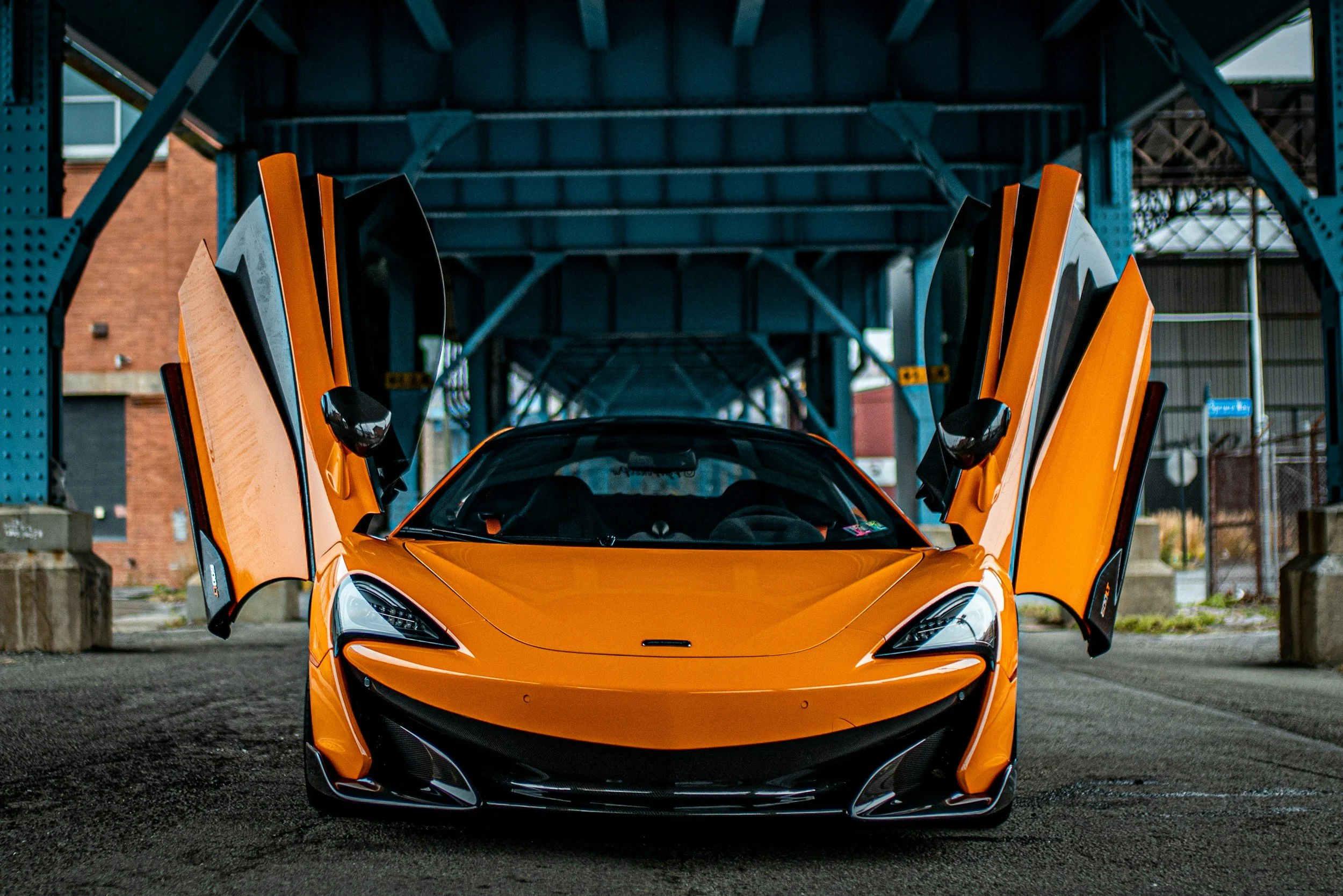 An orange sports car with butterfly doors open under a bridge.