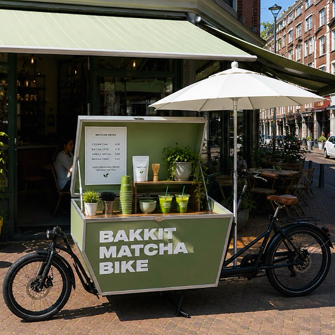 Groene bakfiets met een thee- en matcha-stand in een stad, naast een café met terras, onder een witte parasol.