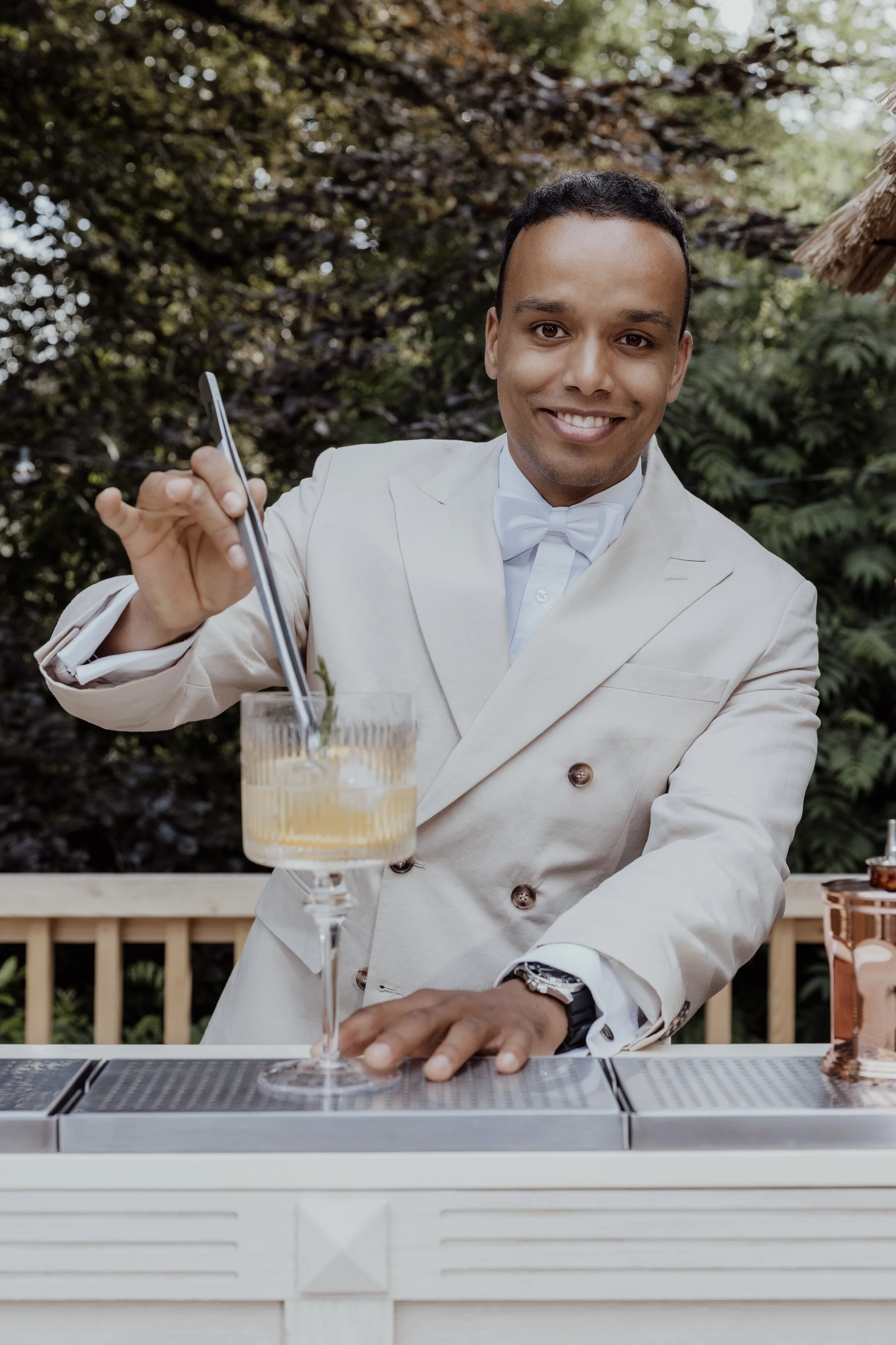 A smiling man in a white tuxedo with a bow tie is making a cocktail outdoors, with greenery in the background.
