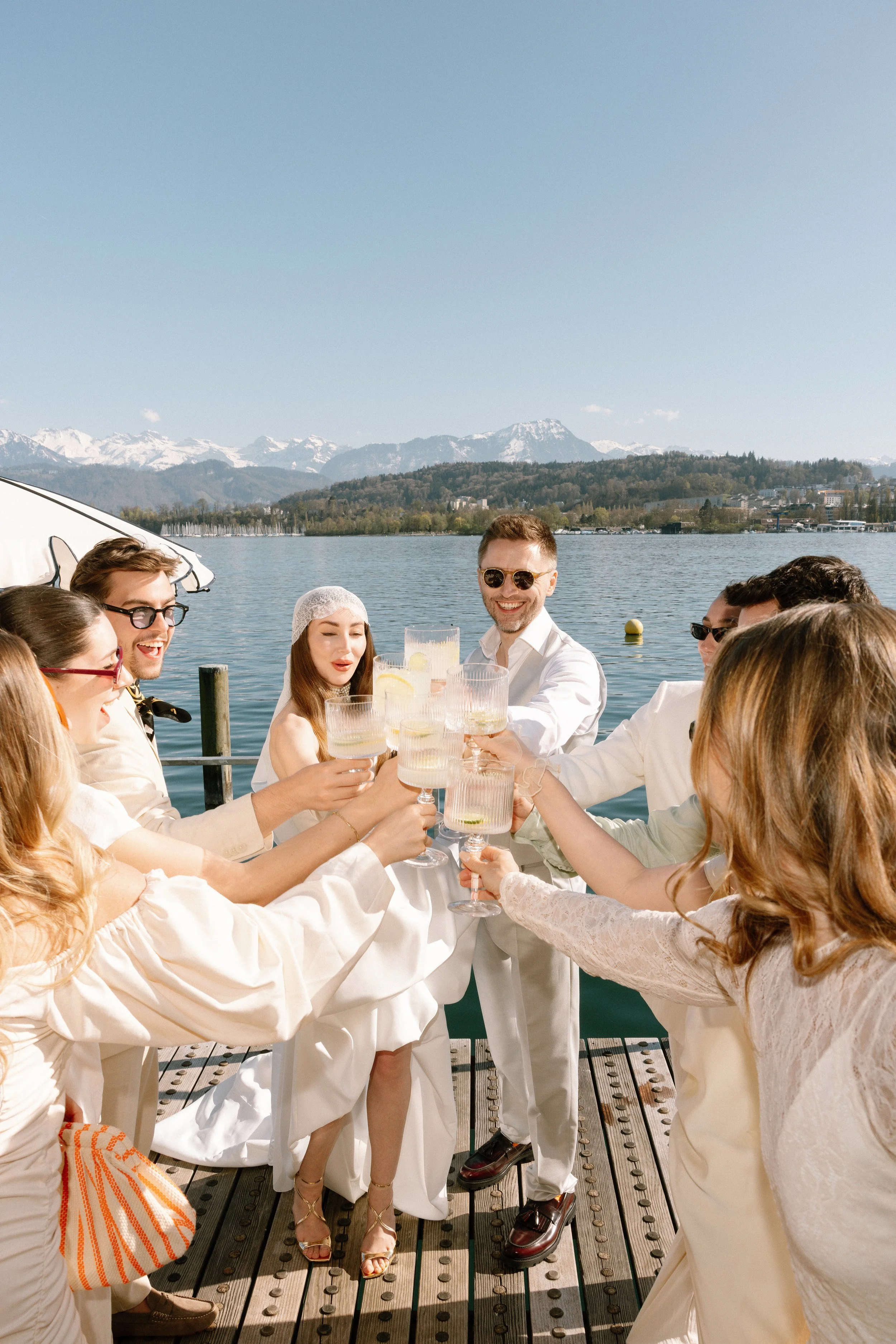 A group of people dressed in white, smiling and holding glasses of drinks, having a toast outdoors on a deck by a lake, with snow-capped mountains in the background.