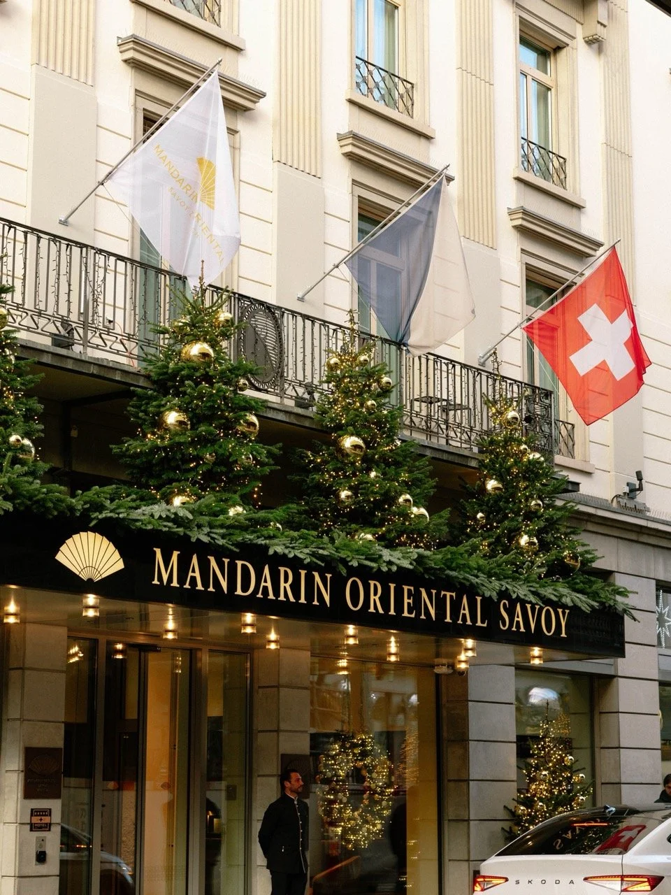 Exterior view of Mandarin Oriental Savoy hotel decorated for Christmas with Christmas trees, gold ornaments, and string lights. Swiss and other flags displayed above the entrance.