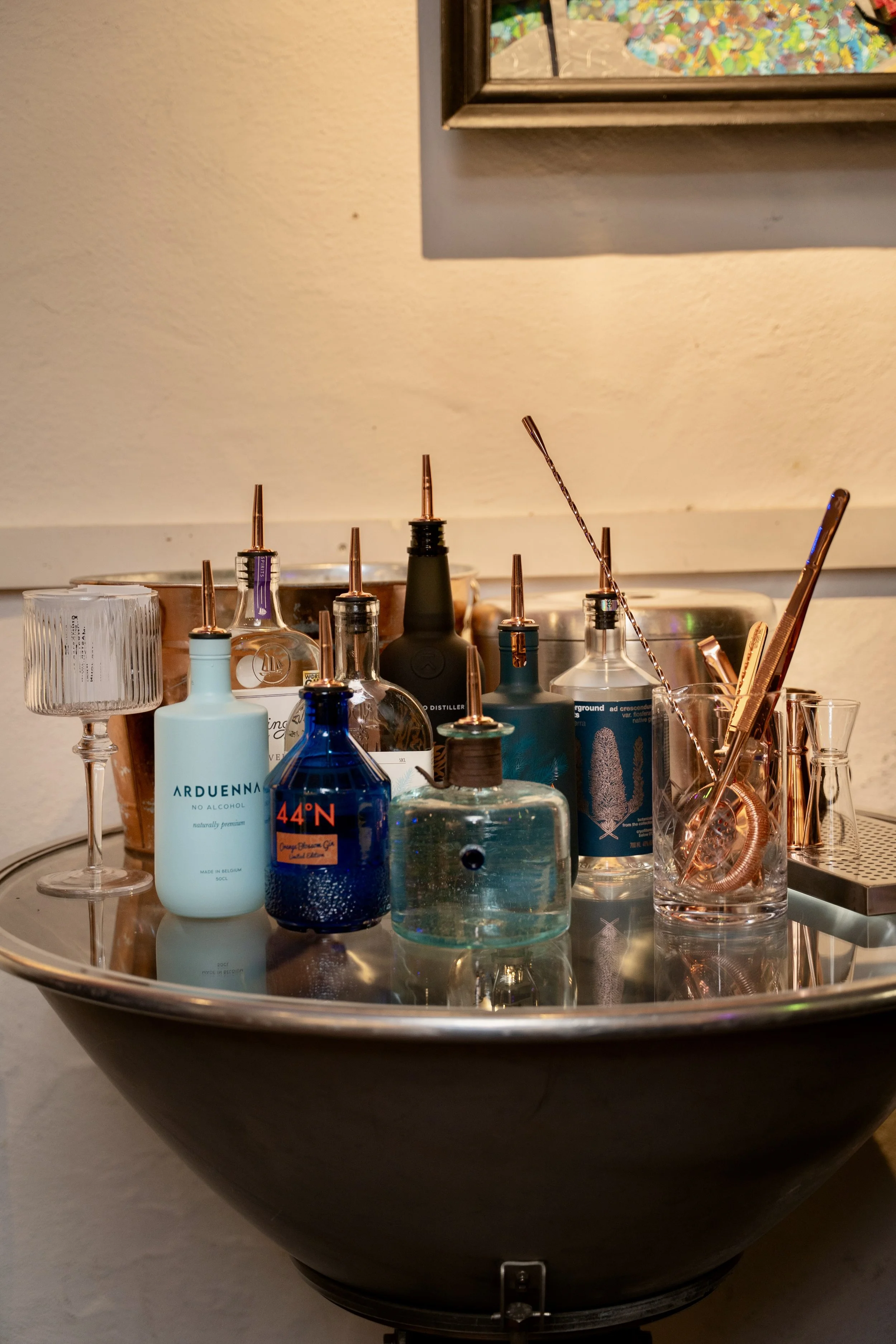 A round, silver tray with various bottles and glassware, including a blue bottle labeled 44°N and a white bottle labeled Arduenna, and some metal straws and utensils. In the background, there is a framed colorful artwork on the wall.