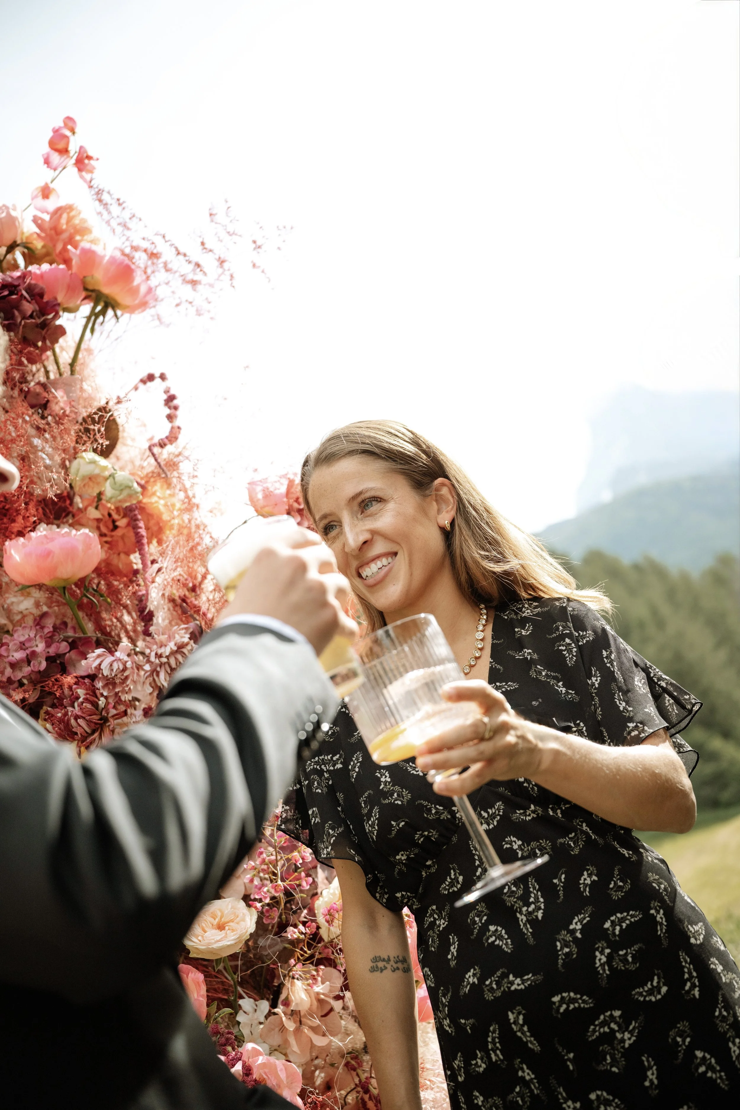 A woman with long hair, smiling, wearing a black dress with white floral patterns, holding a glass of champagne while socializing outdoors during daytime. There is a person in a suit with a striped sleeve offering her a glass, and a backdrop of mount