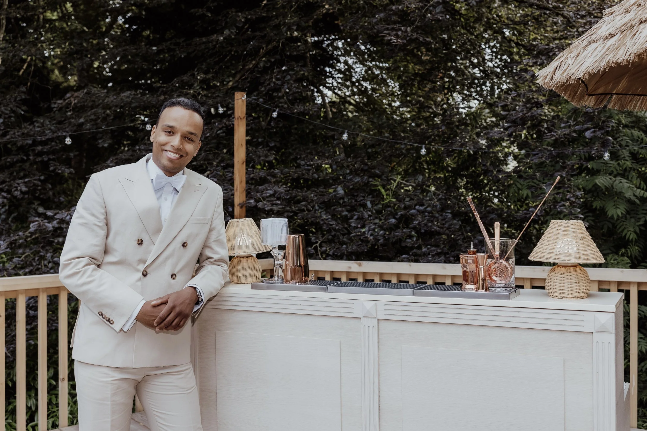 A smiling man in a white tuxedo standing outdoors next to a bar with various cocktail making tools and glasses, set against a wooded background.
