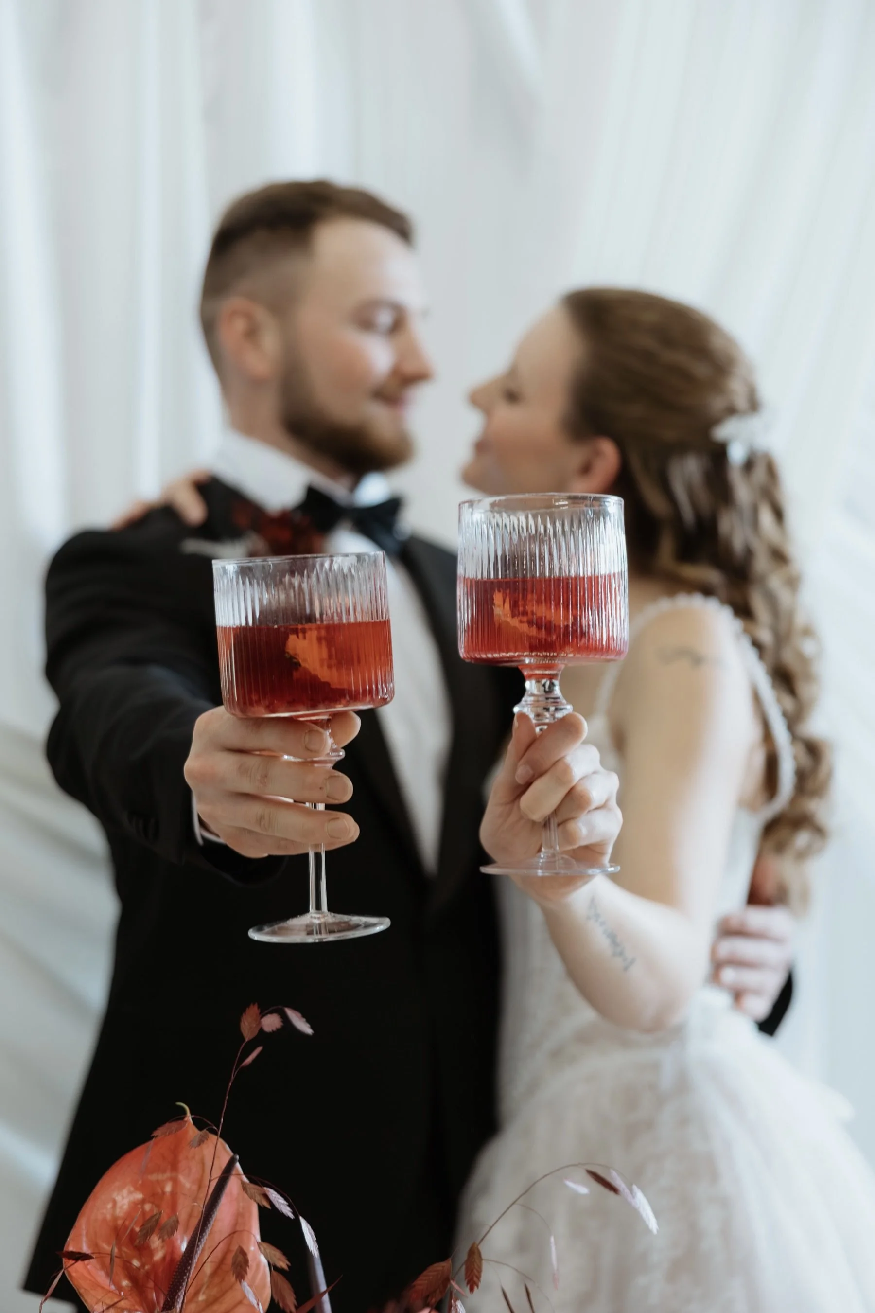A man and a woman dressed in formal wedding attire holding wine glasses with red beverage, smiling and looking at each other, with a blurred background of white curtains.