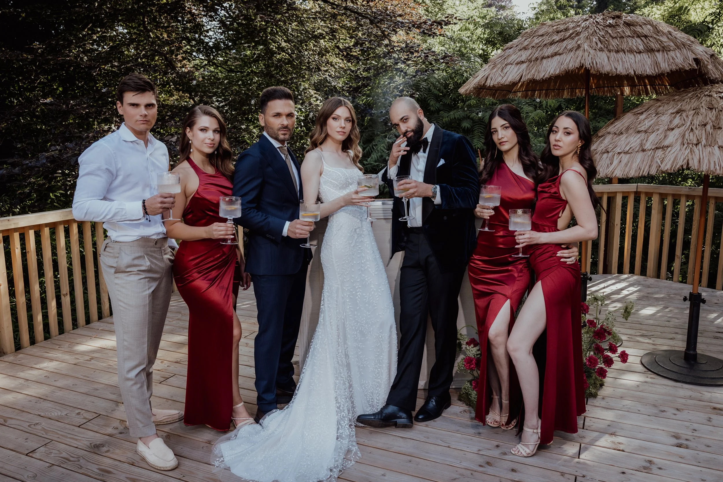 Group of seven people, including bride and groom, holding glasses, standing on a wooden deck outdoors with lush greenery and thatched umbrellas in the background.