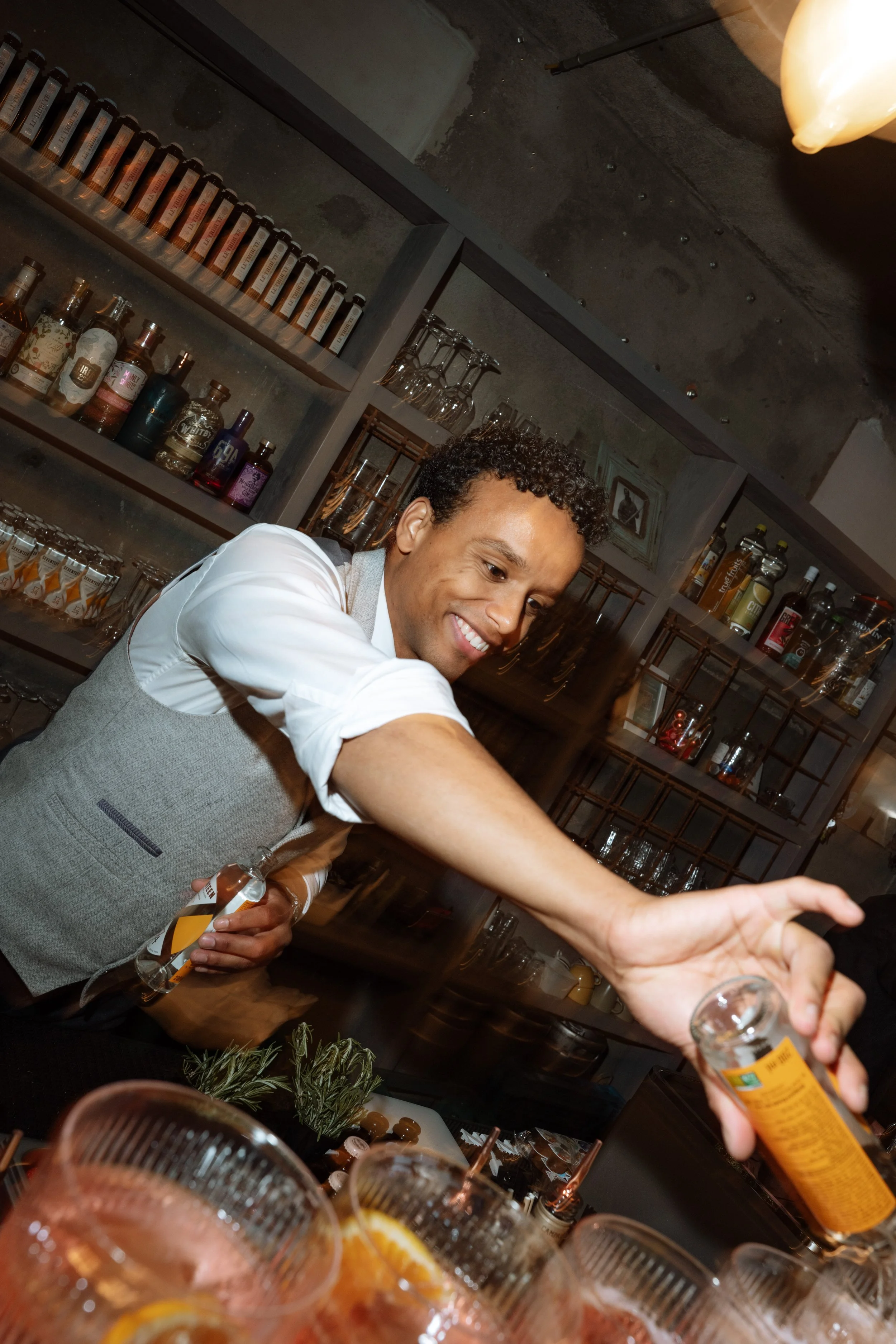A smiling man serving drinks at a bar with liquor bottles and glassware behind him. Several glasses with drinks garnished with orange slices are in the foreground.