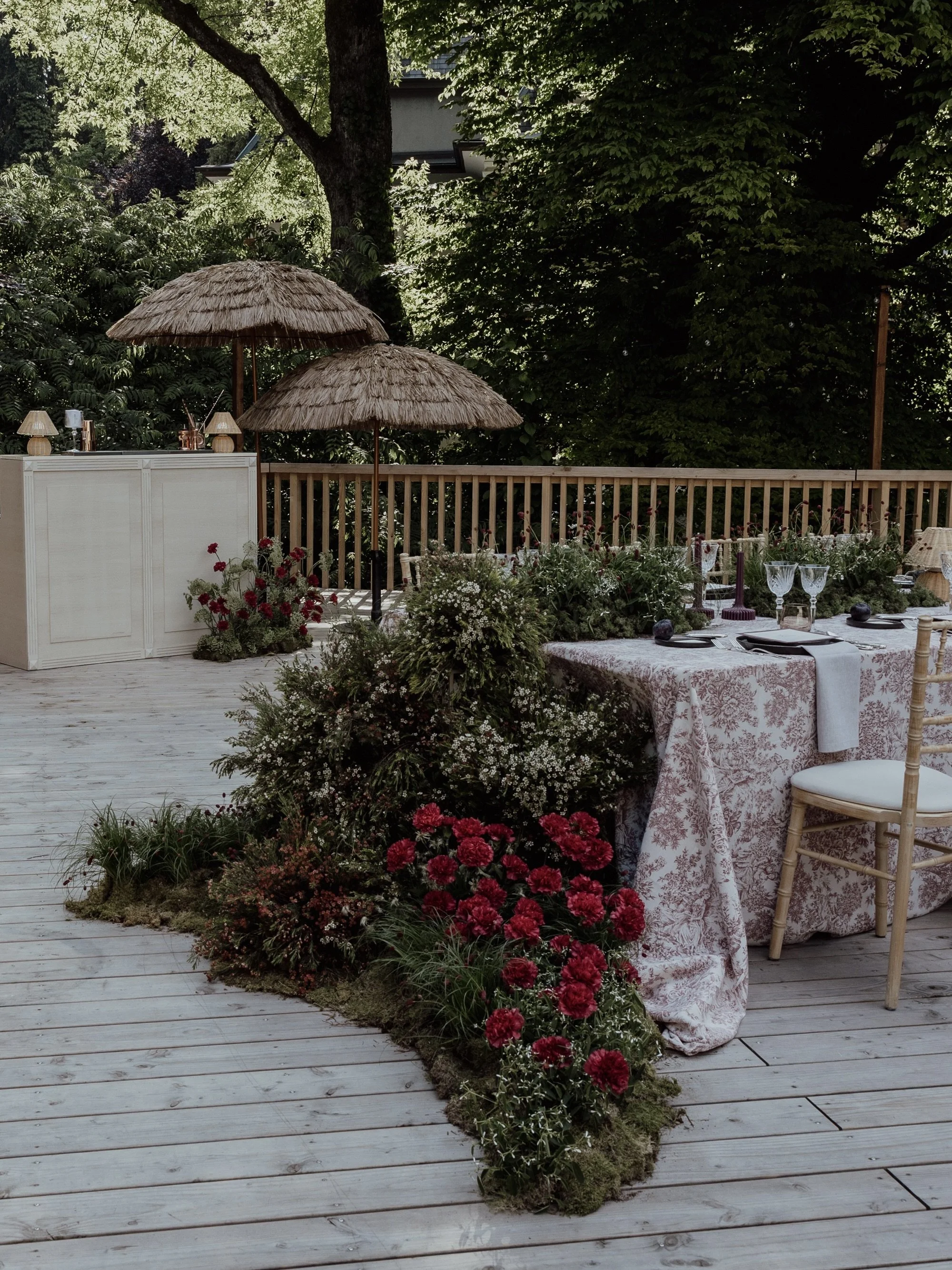 A decorated outdoor dining area on a wooden deck, with floral arrangements, two thatched umbrellas, and glassware on a table with a patterned cloth, surrounded by lush green trees and fencing.