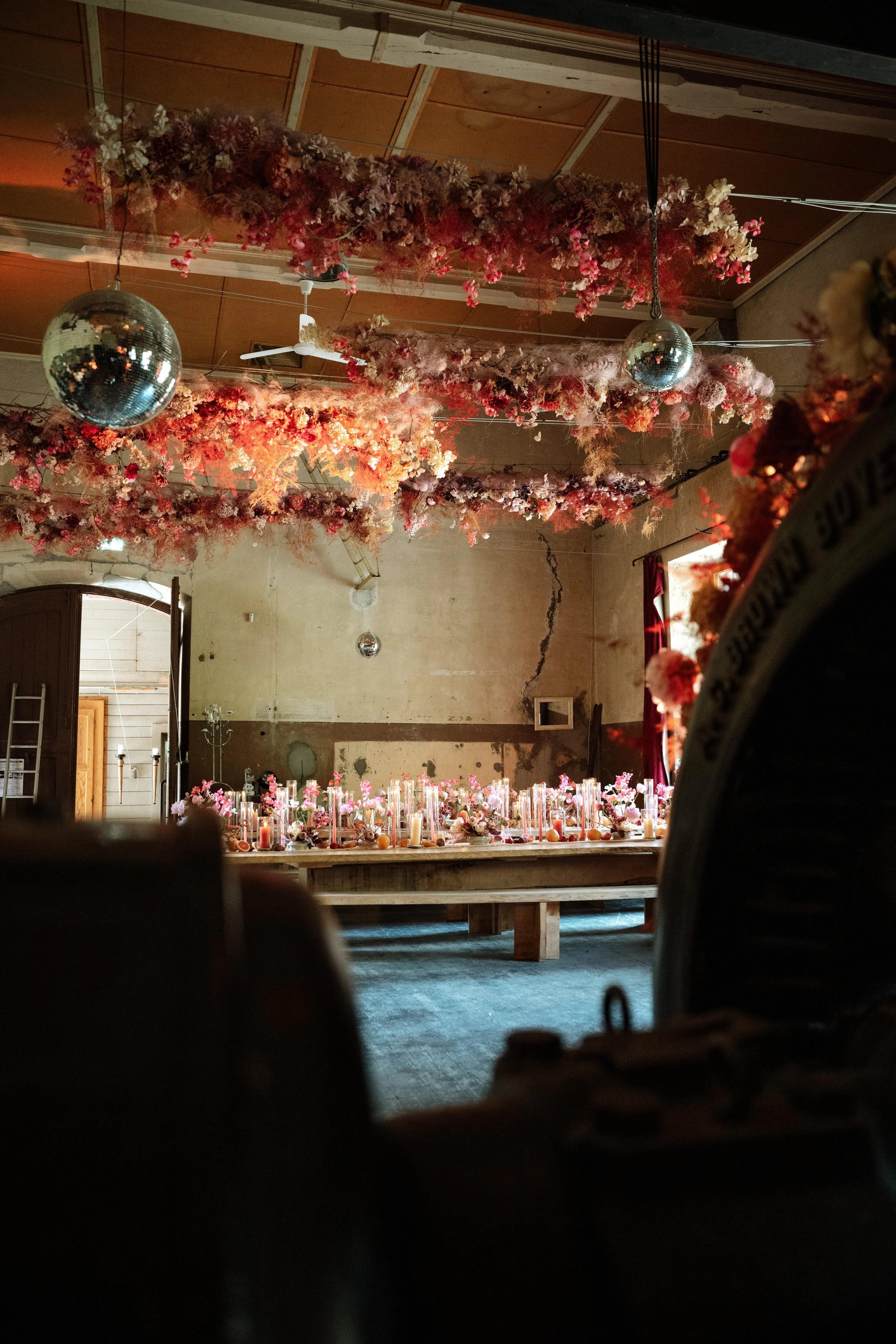 A decorated indoor space with hanging pink floral arrangements and two disco balls on the ceiling. In the background, there is a table with candles and flower decorations, and a cracked wall with a small window. The room has a rustic, vintage atmosph