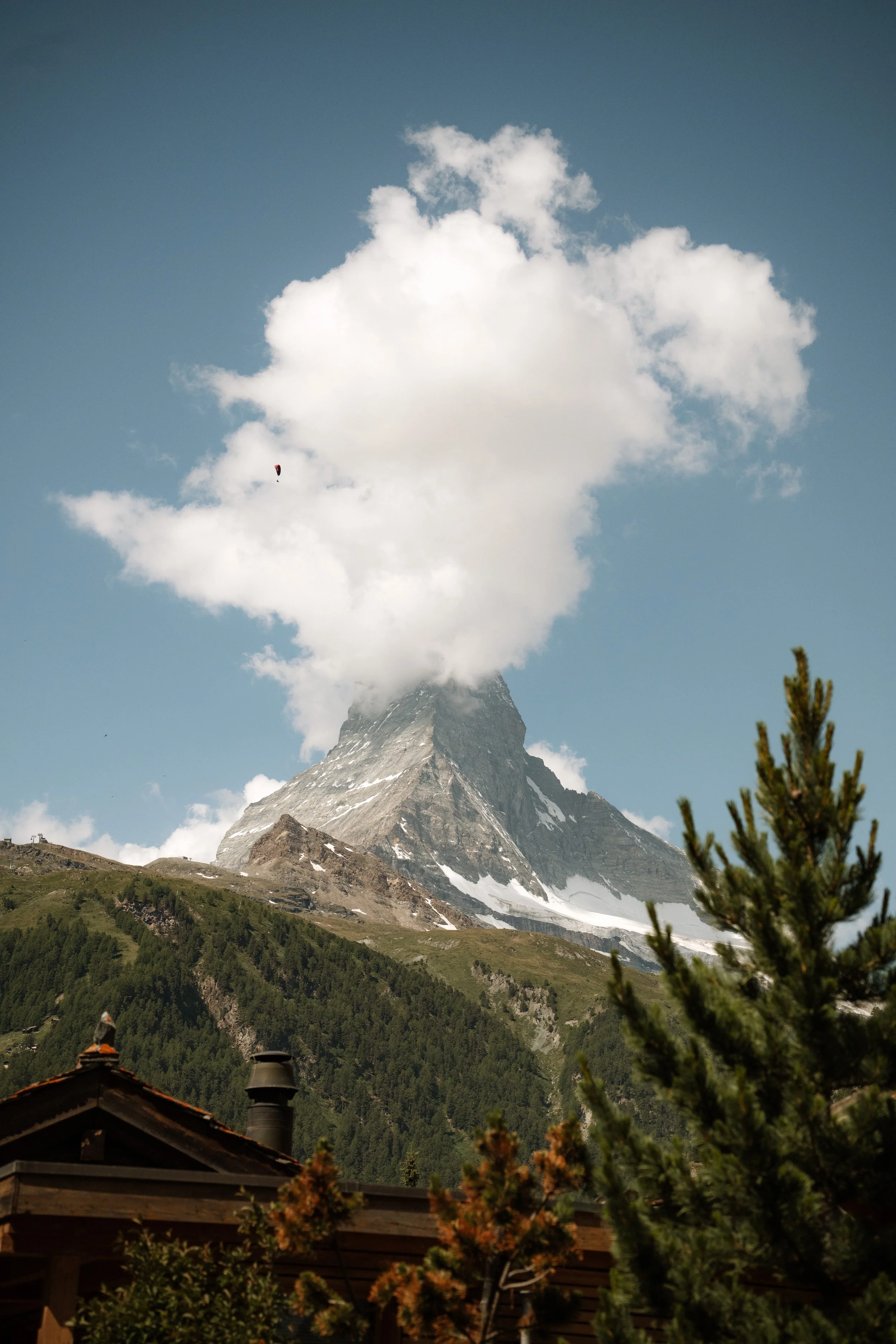 Snow-capped mountain with large cloud formations above it, a paraglider flying near the cloud, and rooftops and trees in the foreground.