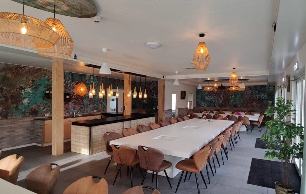 Een vergaderruimte met een grote witte tafel, meerdere stoelen, groene planten, en wanddecoraties, verlicht door hanglampen en plafondverlichting.