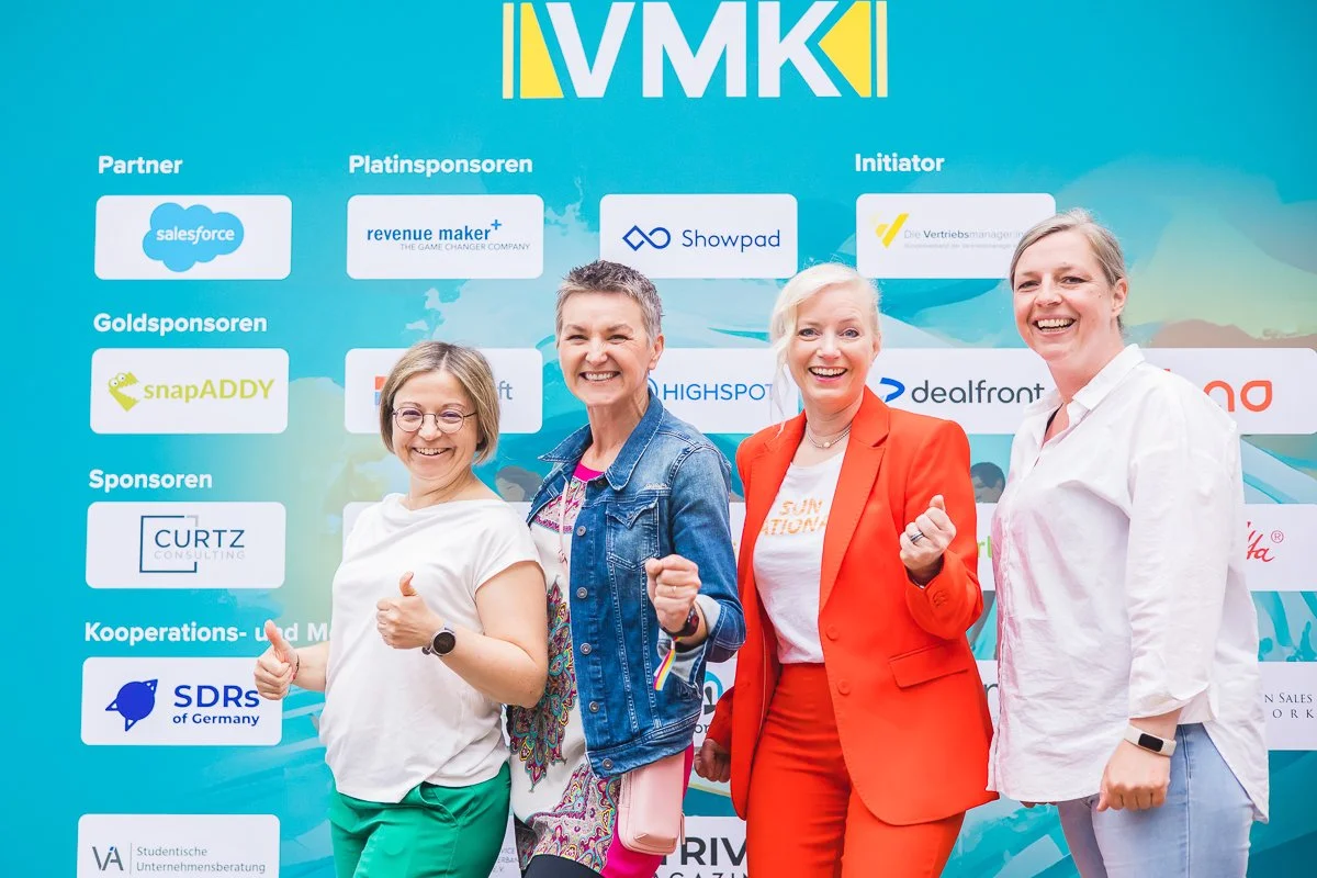 Vier Frauen beim Event, lächelnd, vor einer blauen Veranstaltungswand mit Logos und Text, begrüßen sich und zeigen Handzeichen.