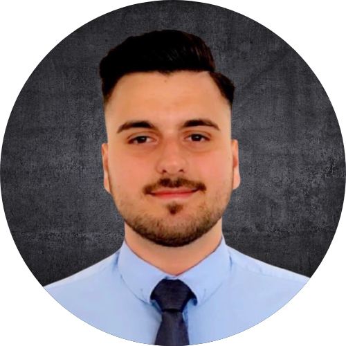 Ein junger Mann mit dunklem Haar, Bart und Schnurrbart trägt einen hellblauen Hemd und eine dunkle Krawatte vor einem schwarzen Hintergrund.