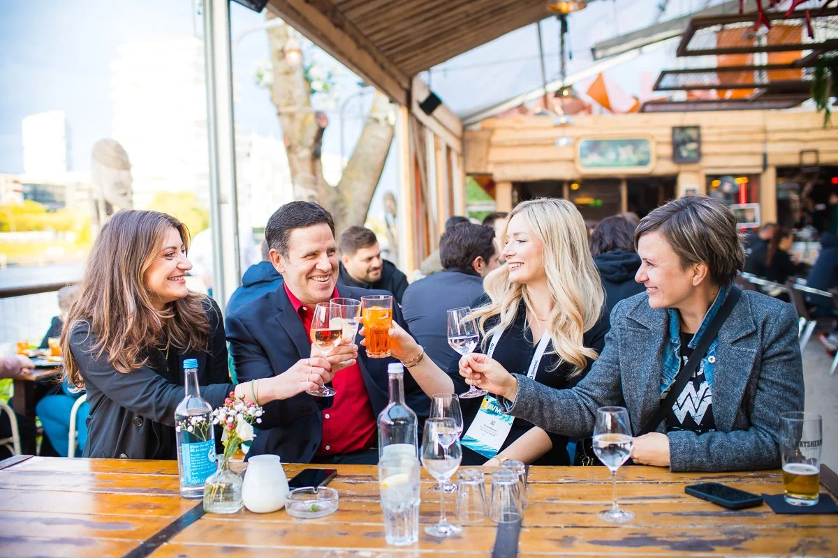 Vier Menschen, zwei Frauen und zwei Männer, sitzen an einem Holztisch in einer gemütlichen Bar oder Restaurant, es wird ein Toast mit verschiedenen Getränken gemacht, umgeben von weiteren Gästen in einem offenen, holzverkleideten Raum.