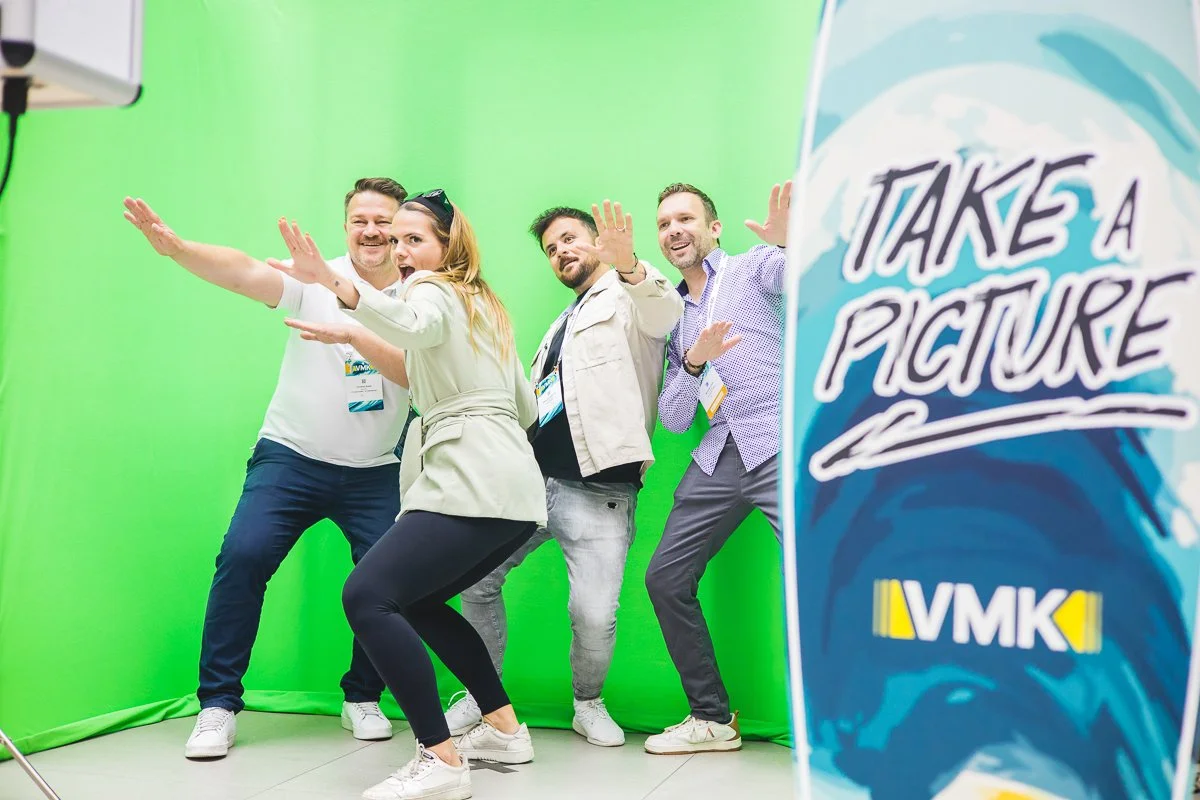 Vier Menschen posieren vor einem grünen Bildschirm und machen eine lustige Pose. Auf der rechten Seite ist ein Banner mit der Aufschrift „Take a Picture“ und dem Logo VMK.