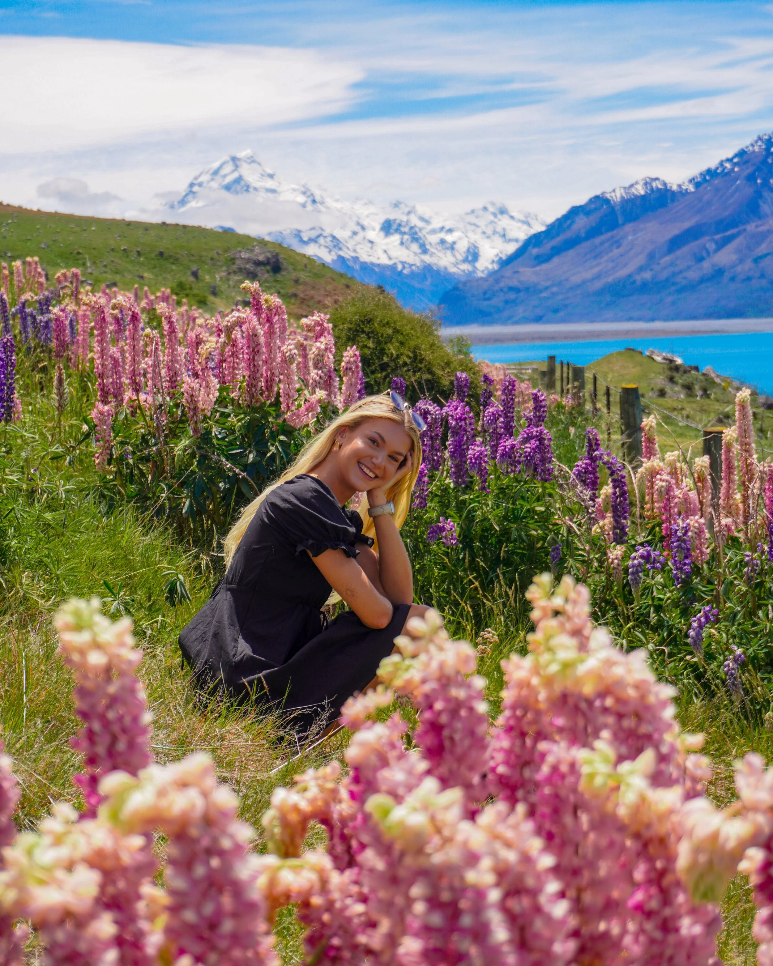 A woman sitting in a field of colorful flowers with snow-capped mountains, a lake, and a partly cloudy sky in the background.
