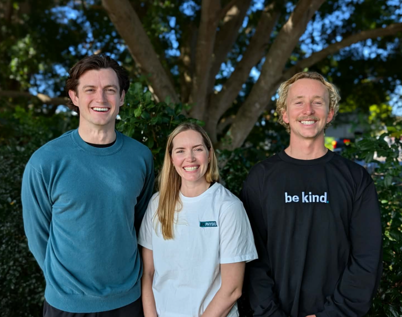The B K Physio team - including Ben, Ellen and Brad - standing outdoors in front of a large tree and greenery and smiling to the camera.