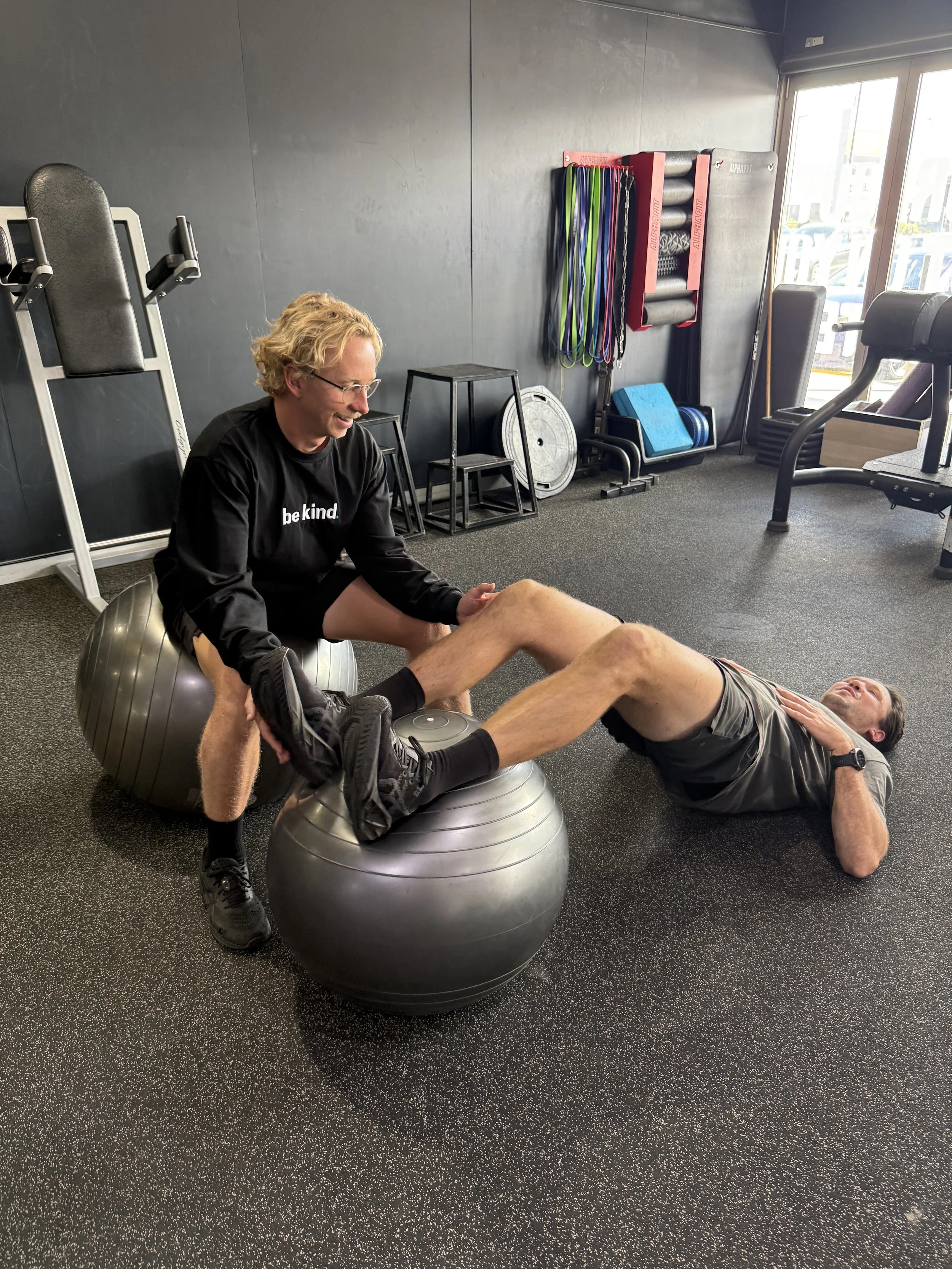 B K Physio physiotherapist is smiling while he assists a man performing leg exercises with an exercise ball in the gym at Jetts Gym Ballina.