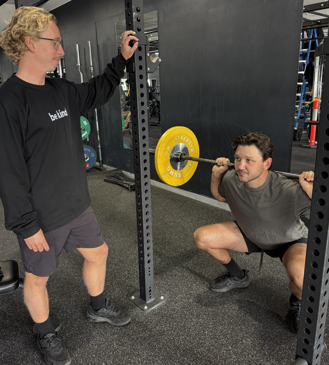 A man squatting with a barbell on his shoulders, lifting weights, while a physiotherapist supervises him in a gym.