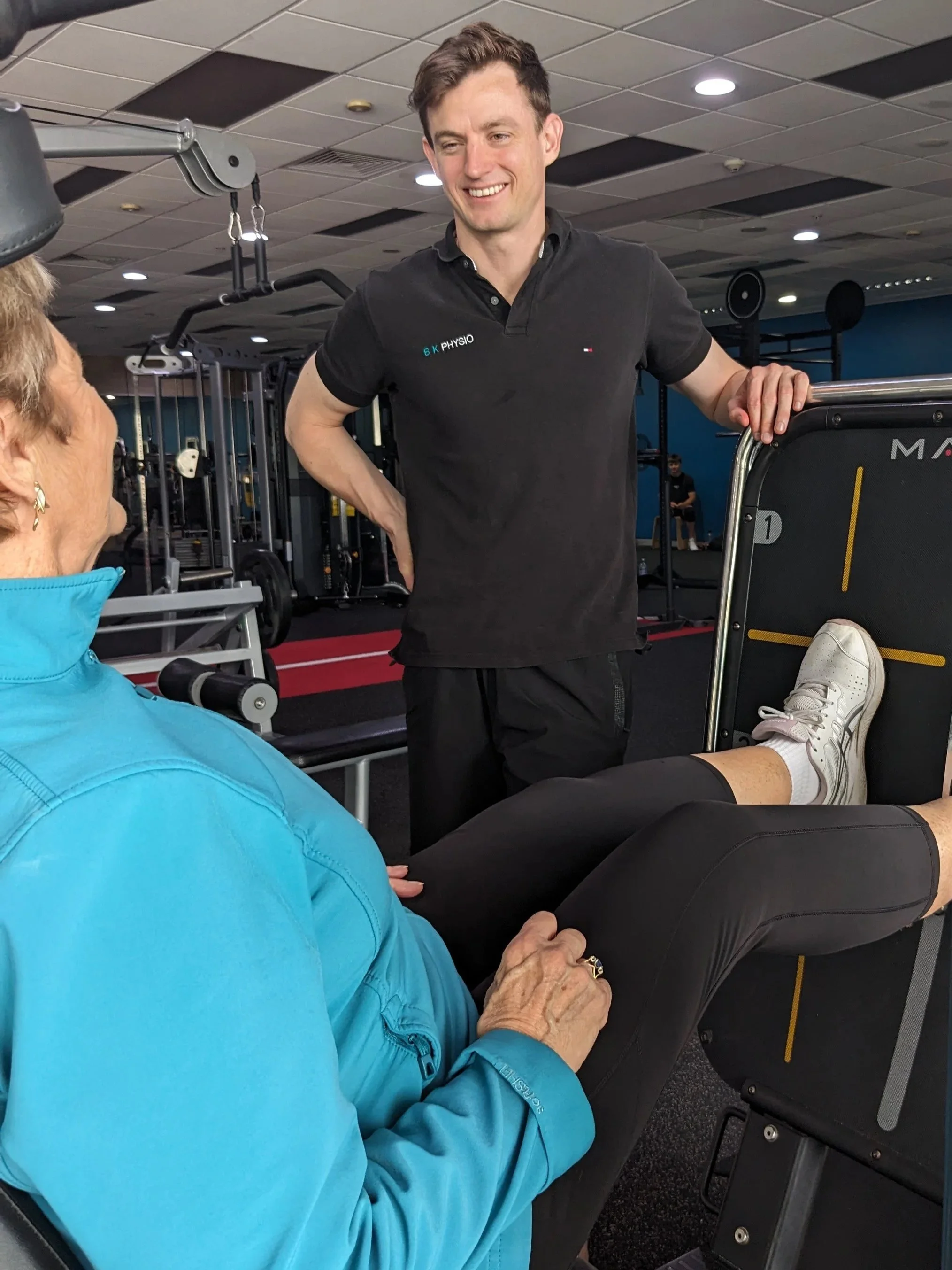 A physiotherapist, is smiling and talking to an elderly woman seated in the gym at Jetts Gym Ballina. The elderly woman is wearing a blue jacket, black pants, and white sneakers. The gym has various fitness equipment in the background.