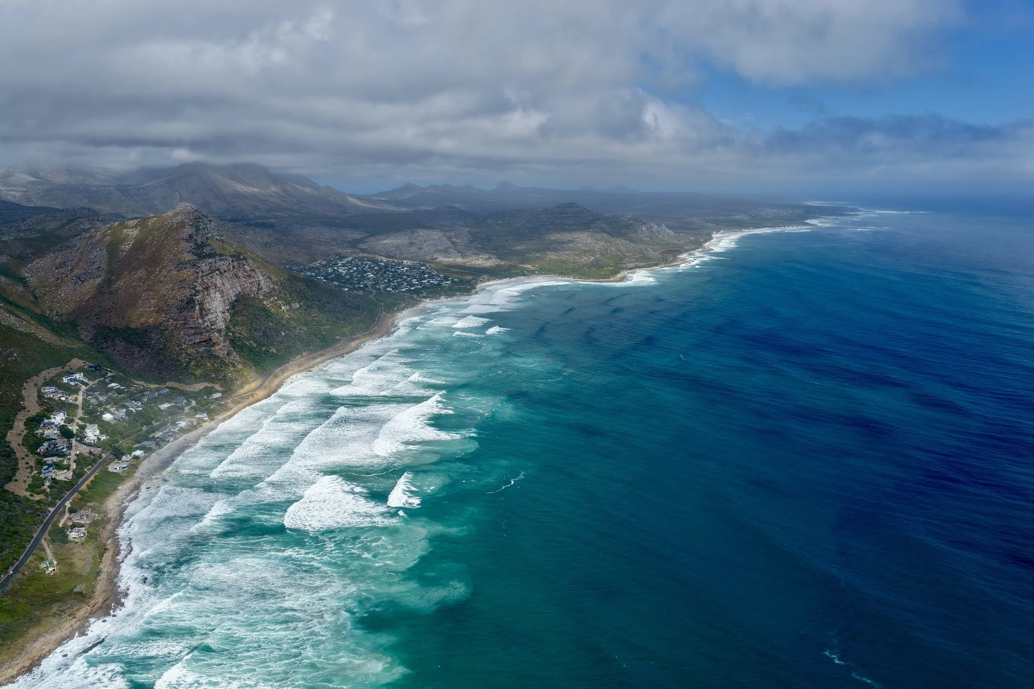 Scarborough, Cape Town aerial view of a rugged coastline with waves crashing onto the shore, green hills, and mountains under cloudy skies.
