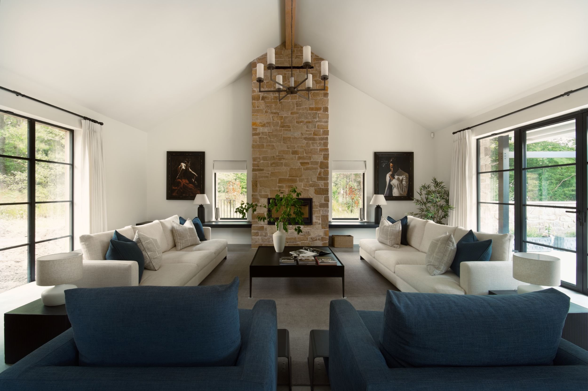 Spacious living room with two large white sofas, two blue armchairs, a stone fireplace in the center, and large windows with white curtains, natural light, and indoor plants.