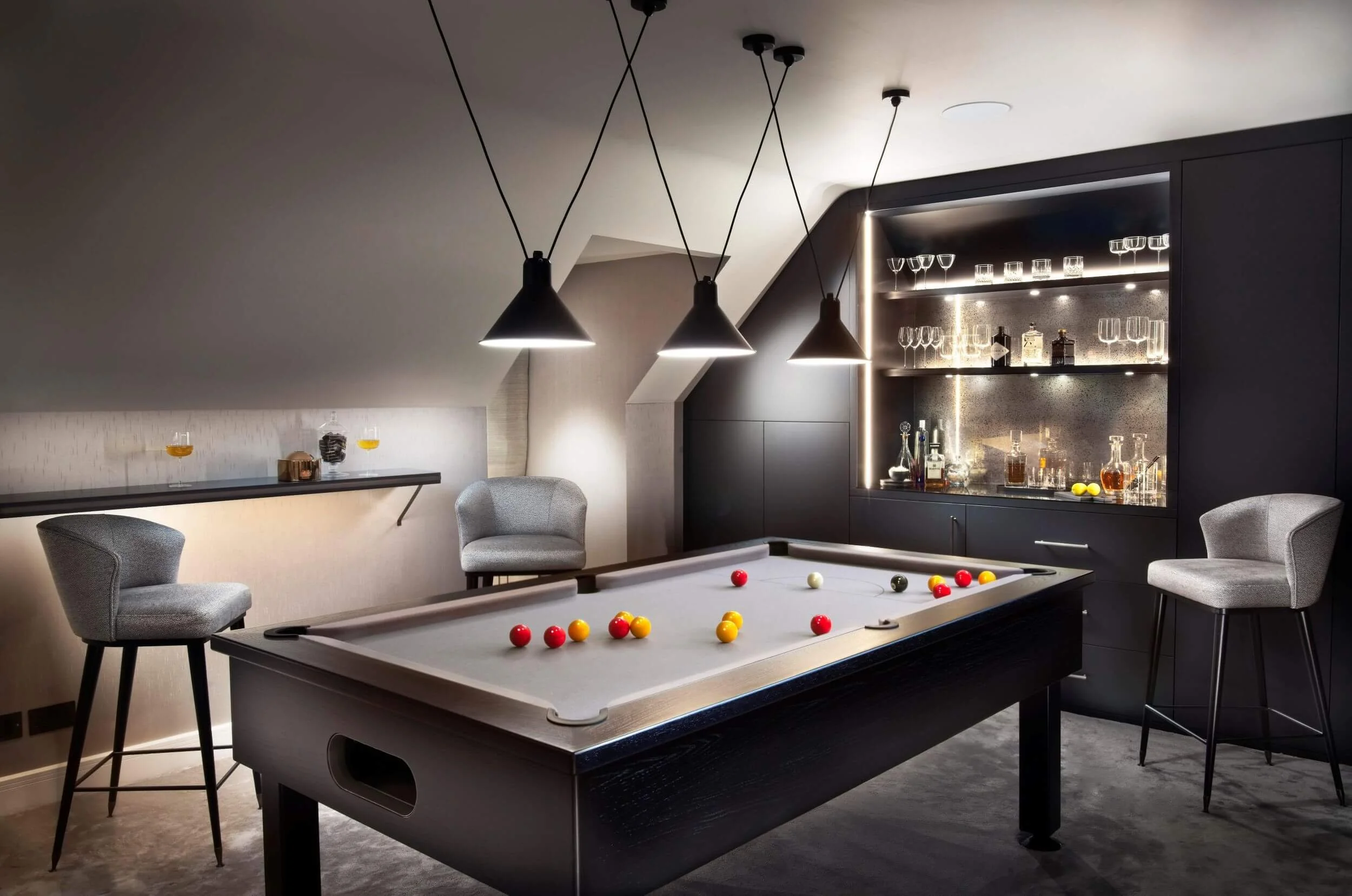 A modern game room with a pool table, gray chairs, and a bar cabinet with glassware and bottles on illuminated shelves.