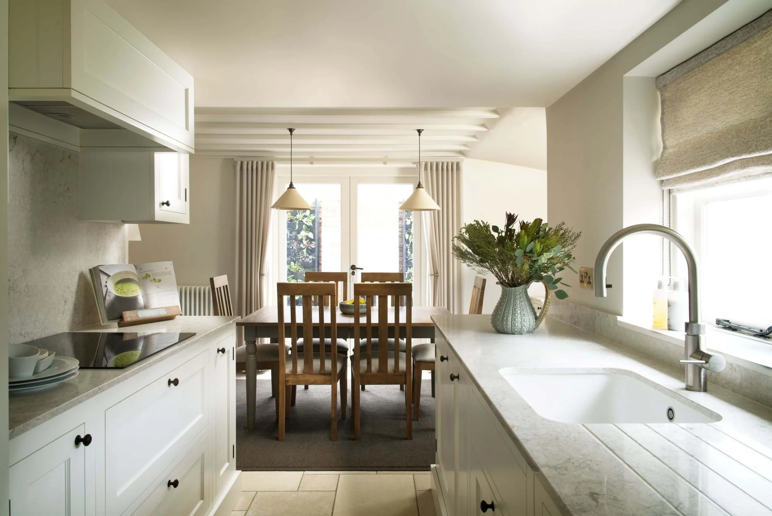 View of a well-lit kitchen overlooking a dining area with wooden table and chairs, decorated with a vase of flowers, with curtains and glass double doors leading to an outside garden.