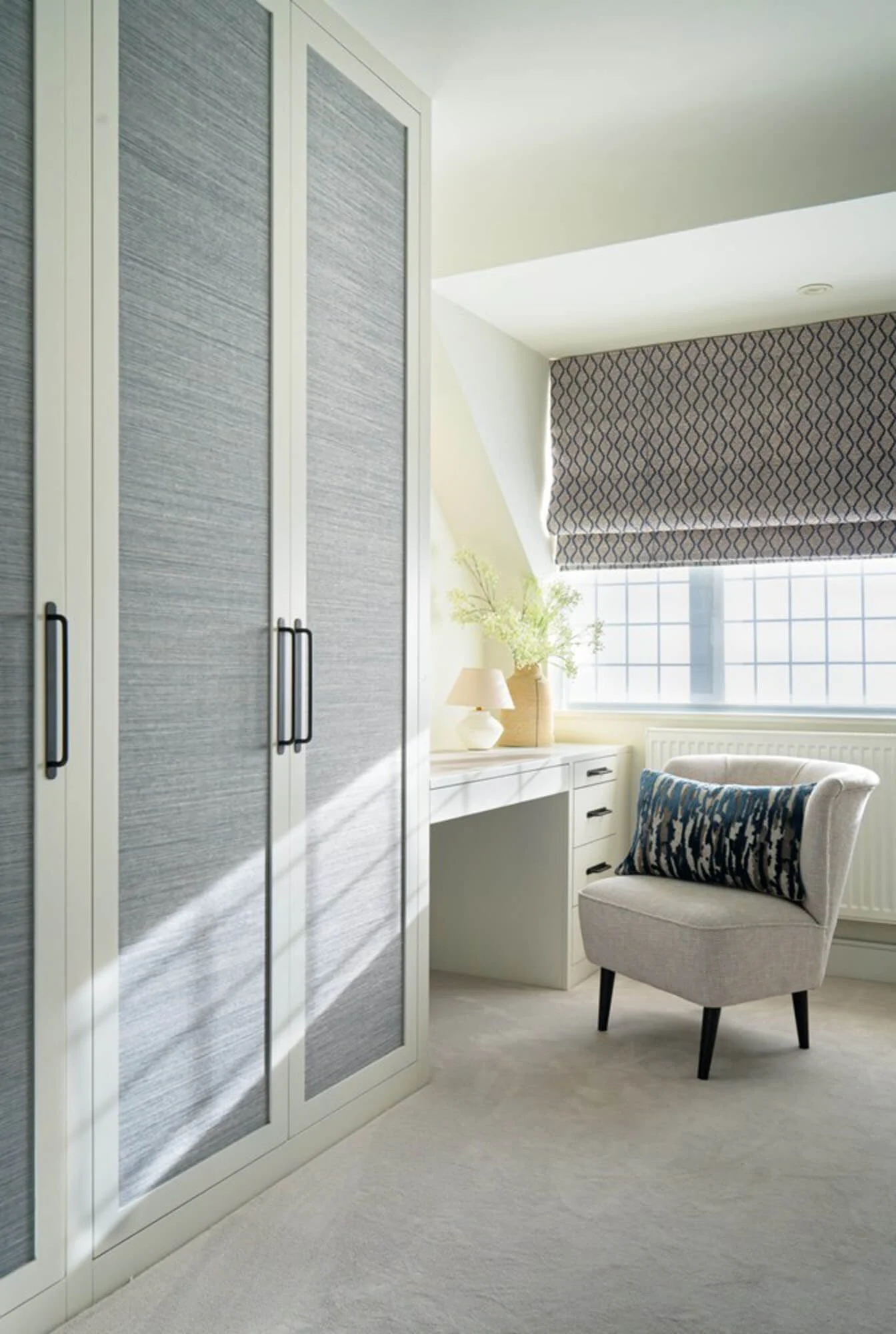 A bright corner of a room with a large window with patterned curtains, a white cabinet with a lamp and vase with flowers, an upholstered chair with a decorative pillow, and a large wardrobe with textured doors.