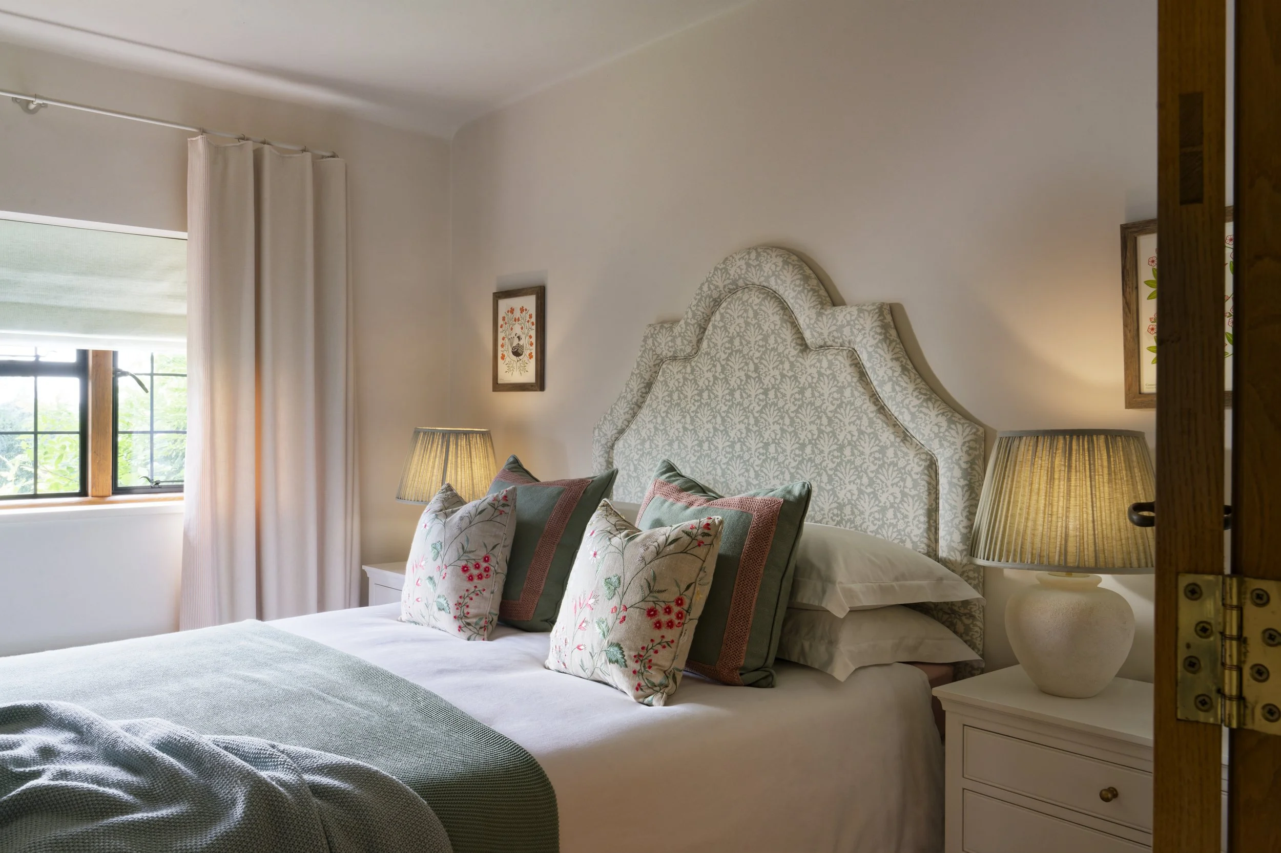 A neatly made bed with white and navy blue bedding, two lamps on white nightstands, a green potted plant, a window with blue trim, and a skylight in a bright bedroom with wooden floors and a beige rug.