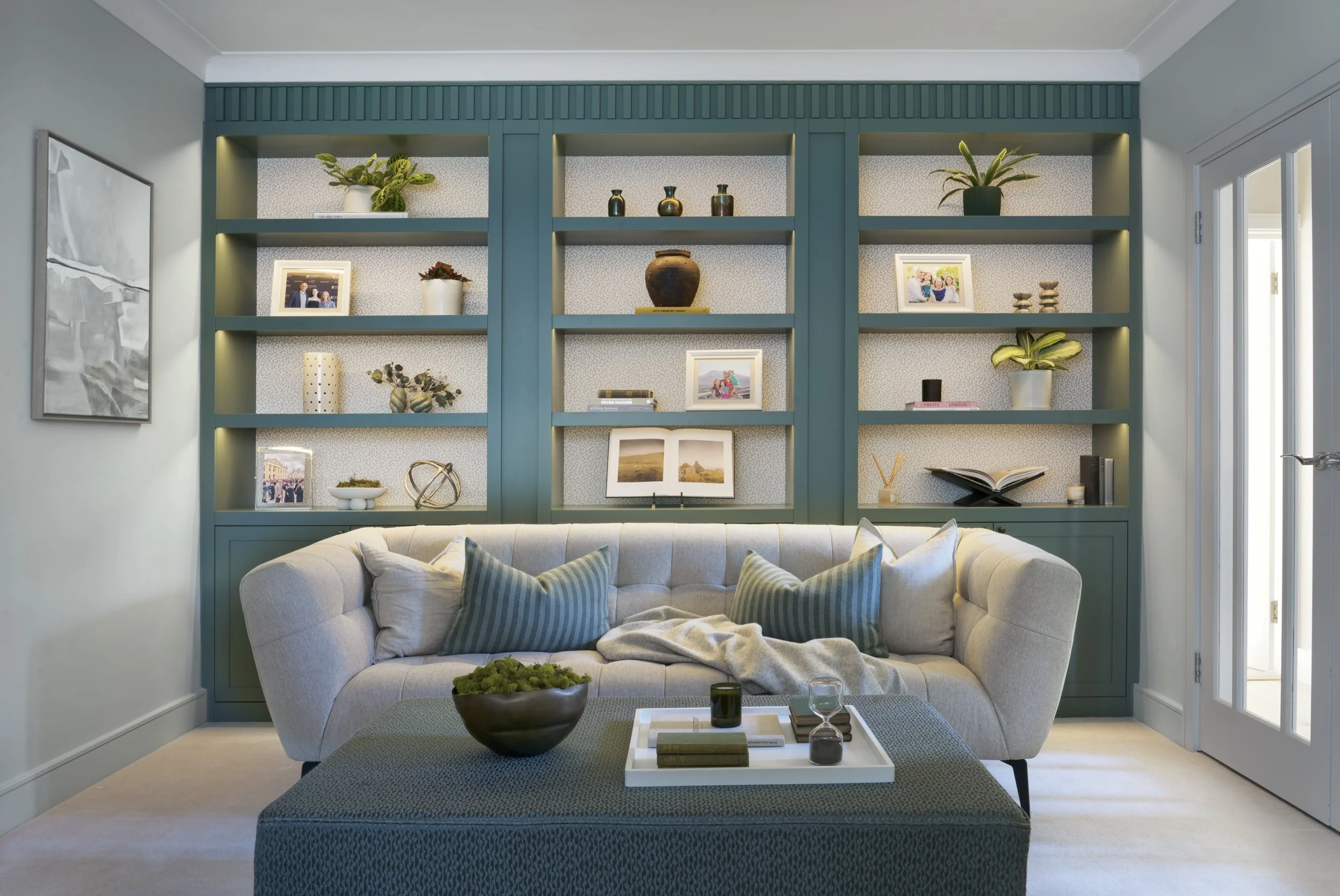 A bright corner of a room with a large window with patterned curtains, a white cabinet with a lamp and vase with flowers, an upholstered chair with a decorative pillow, and a large wardrobe with textured doors.