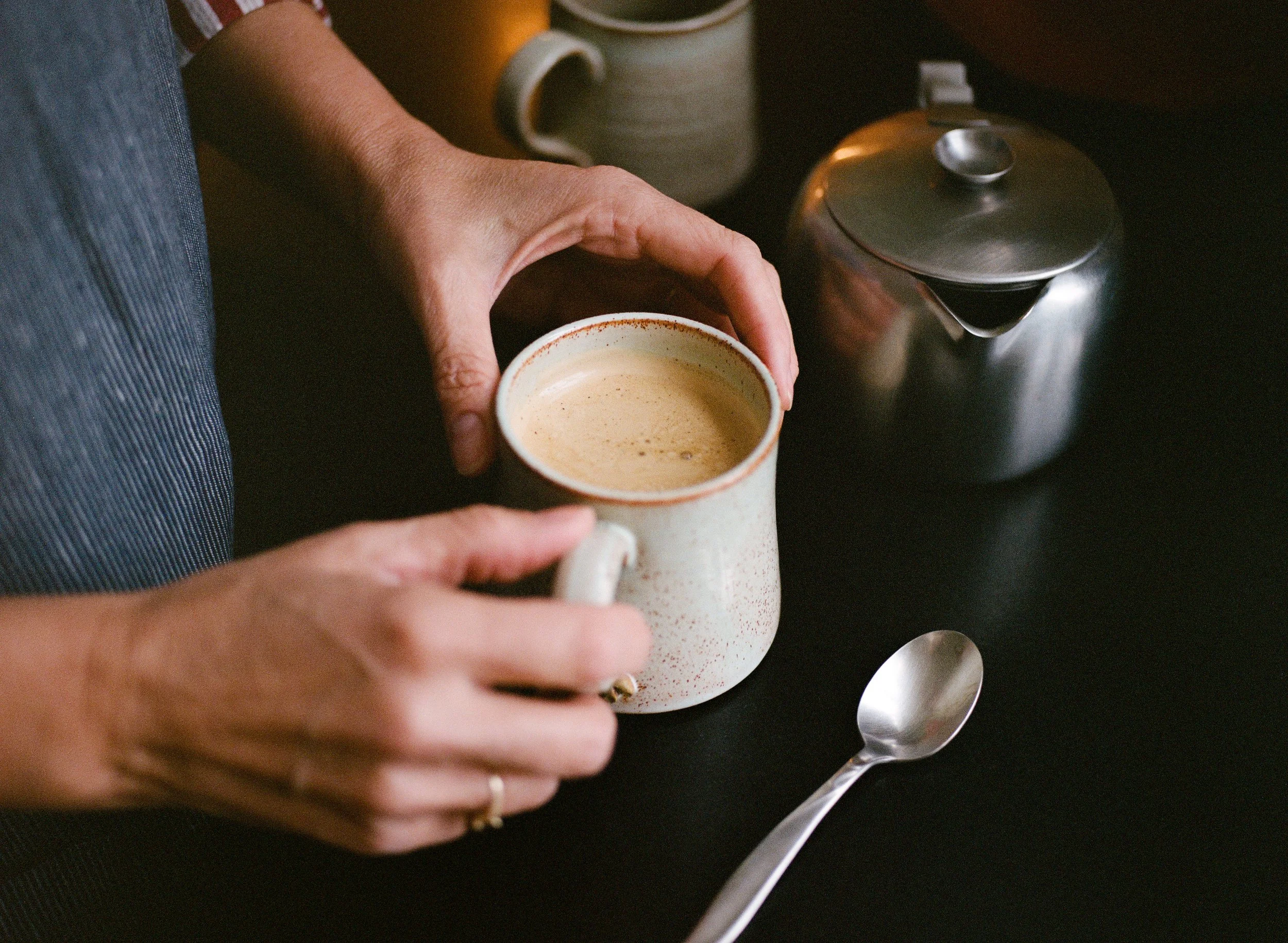 Person holding a mug of coffee with a spoon on a black surface, a stainless steel coffee pot, and other cups in the background.